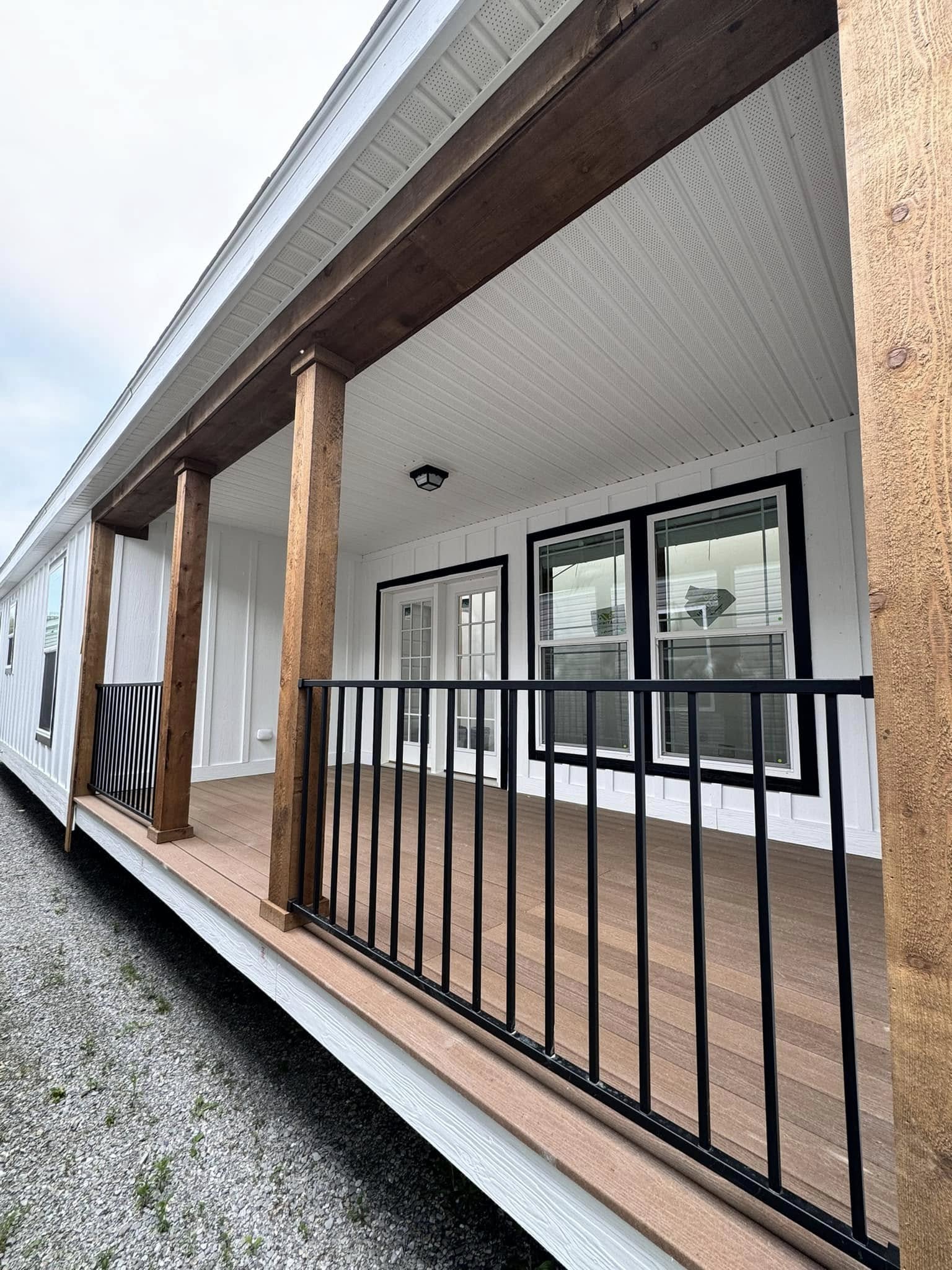 Covered wooden porch with dark metal railing, white siding, and large windows. Wooden posts support the roof, creating a warm, welcoming atmosphere.