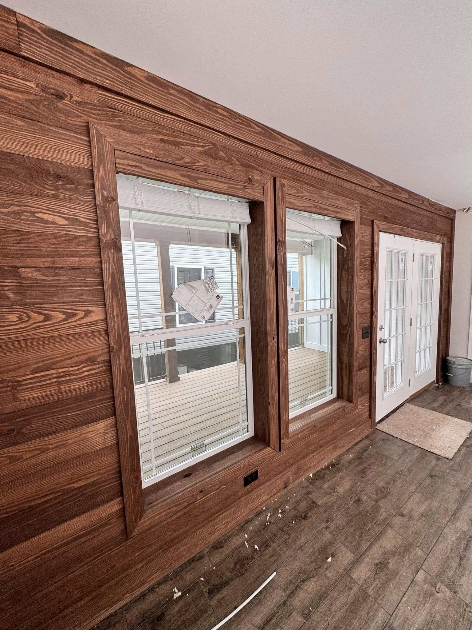 Wood-paneled room with two large windows and double French doors. Sunlight streams through, casting reflections. The floor is wooden, with some debris. The space feels rustic yet bright.