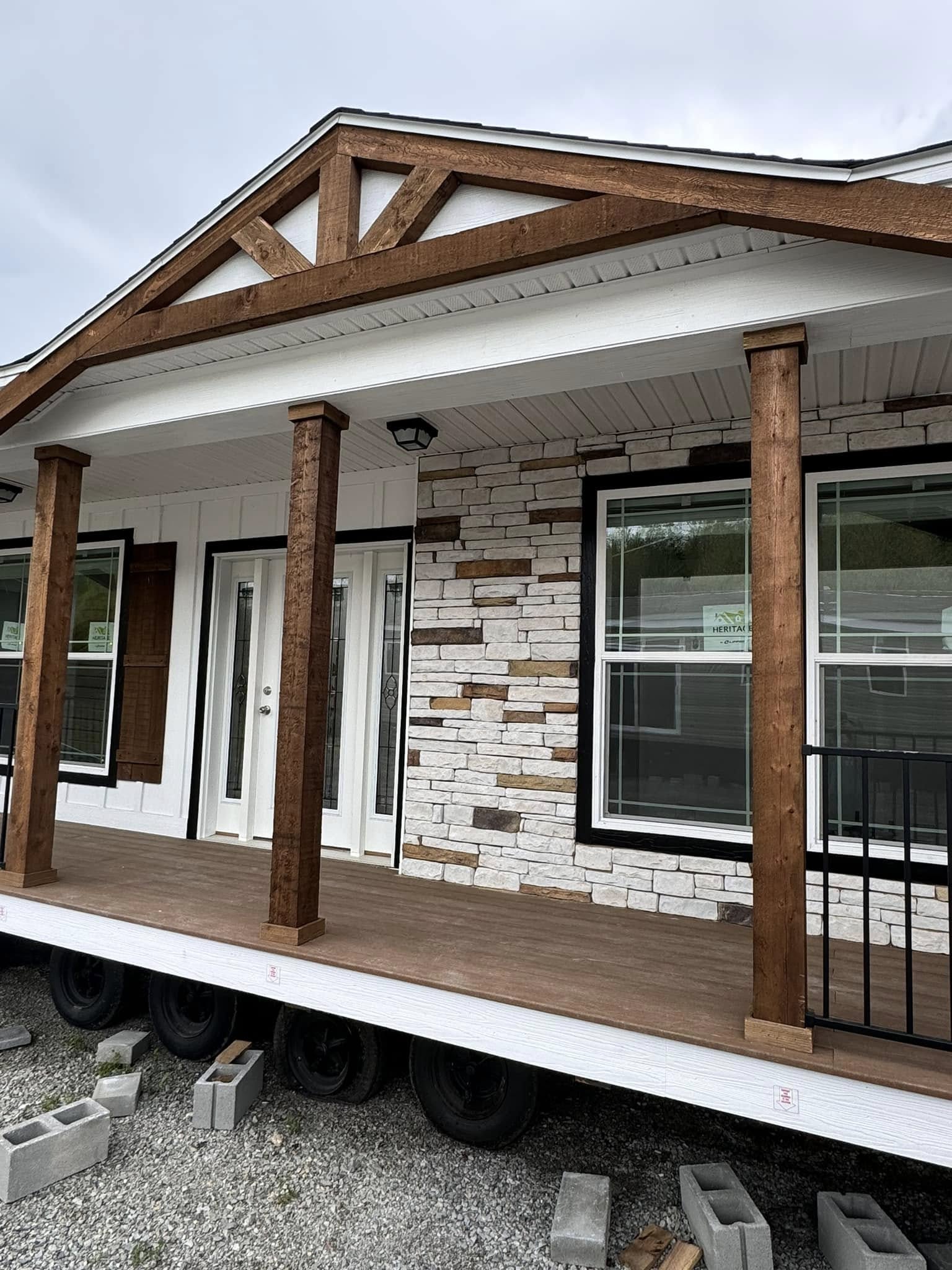 A partially constructed manufactured home with a stone facade and wooden porch. The porch is elevated on wheels, with cinder blocks nearby. Cloudy sky overhead.