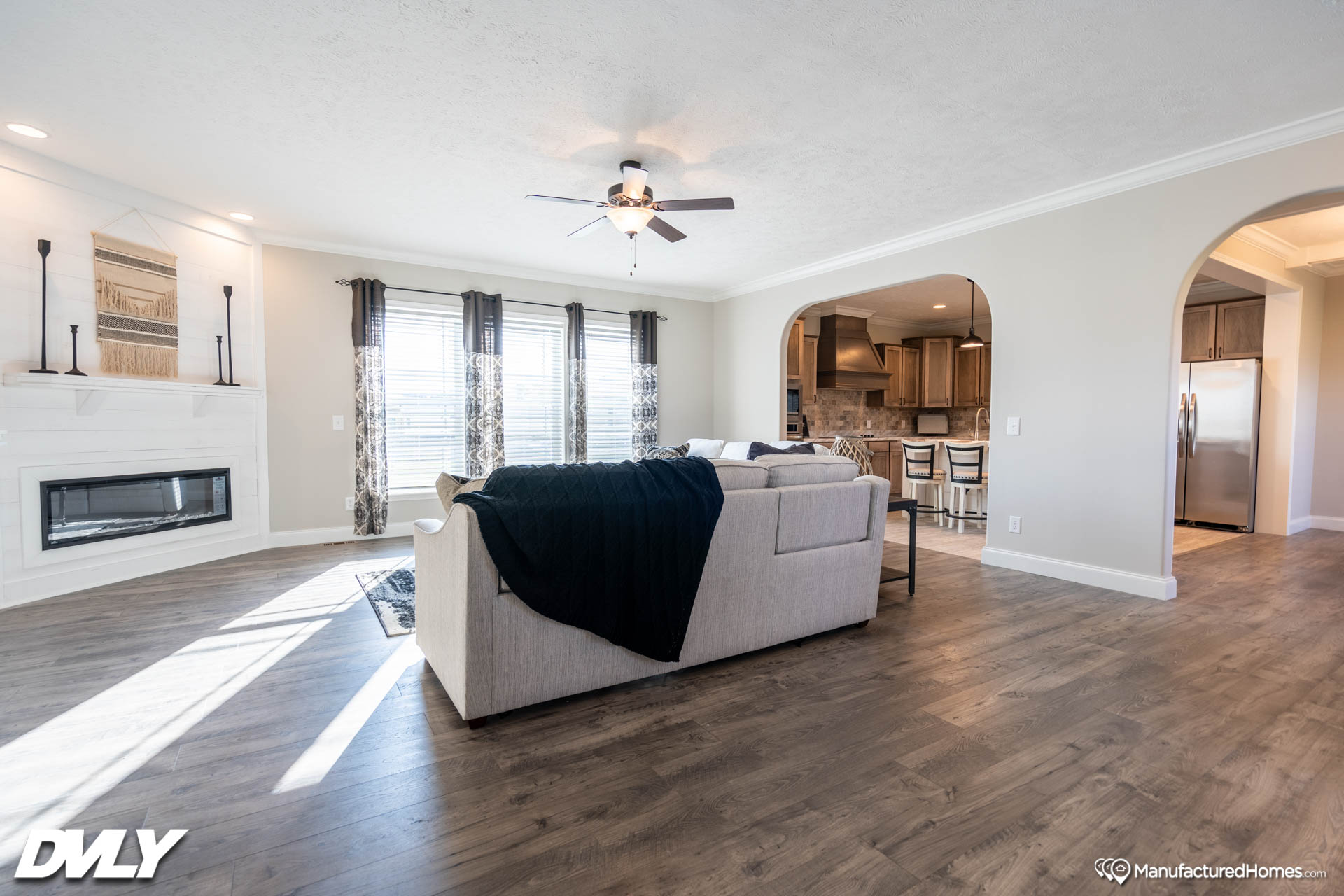 Spacious living room with light wood flooring, a cream-colored sofa, and a dark throw. Sunlight streams through patterned curtains. A ceiling fan hangs overhead, and an archway leads to a modern kitchen. The atmosphere is bright and airy.