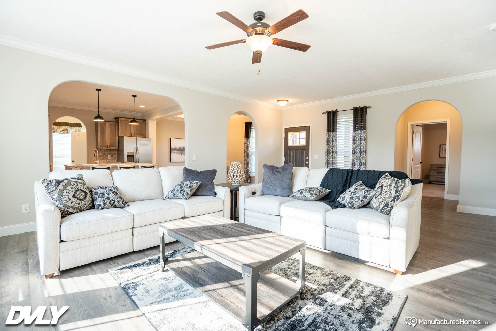 Bright living room with two white sofas featuring patterned pillows, a wooden coffee table on a patterned rug, and a ceiling fan. Arched doorways lead to a kitchen and hallway. Natural light fills the space, creating a cozy, inviting atmosphere.