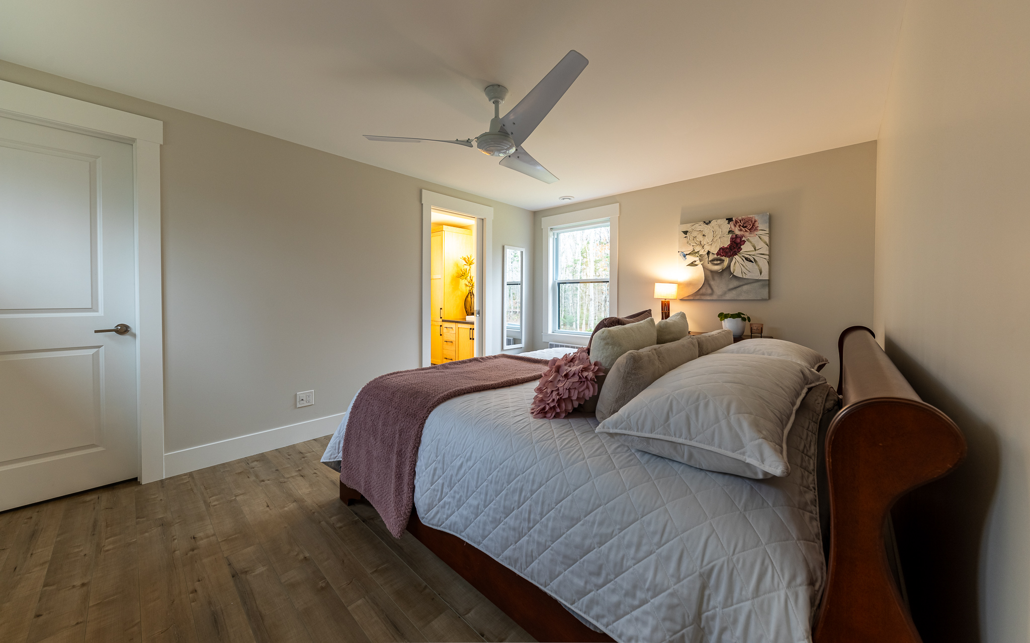 A cozy bedroom with a neatly made bed featuring white and mauve bedding. Soft lighting creates a calm ambiance. Open door shows a lit bathroom.