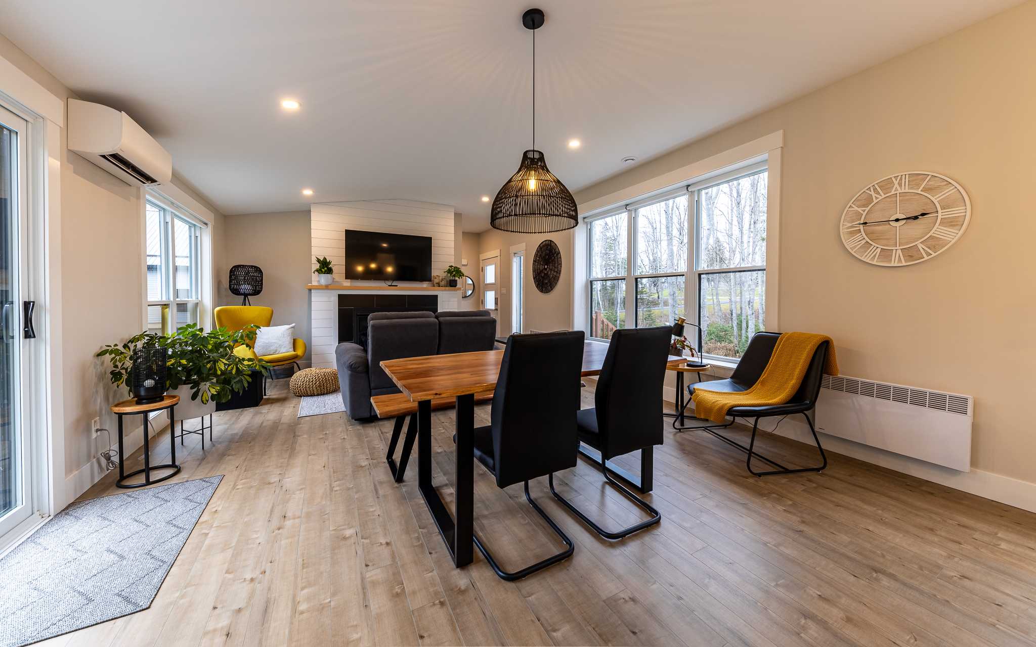 Modern living-dining room with beige walls, wood flooring, and large windows. Features include a wooden dining table, black chairs, yellow armchairs, a wall clock, and a hanging light. Cozy and inviting atmosphere.