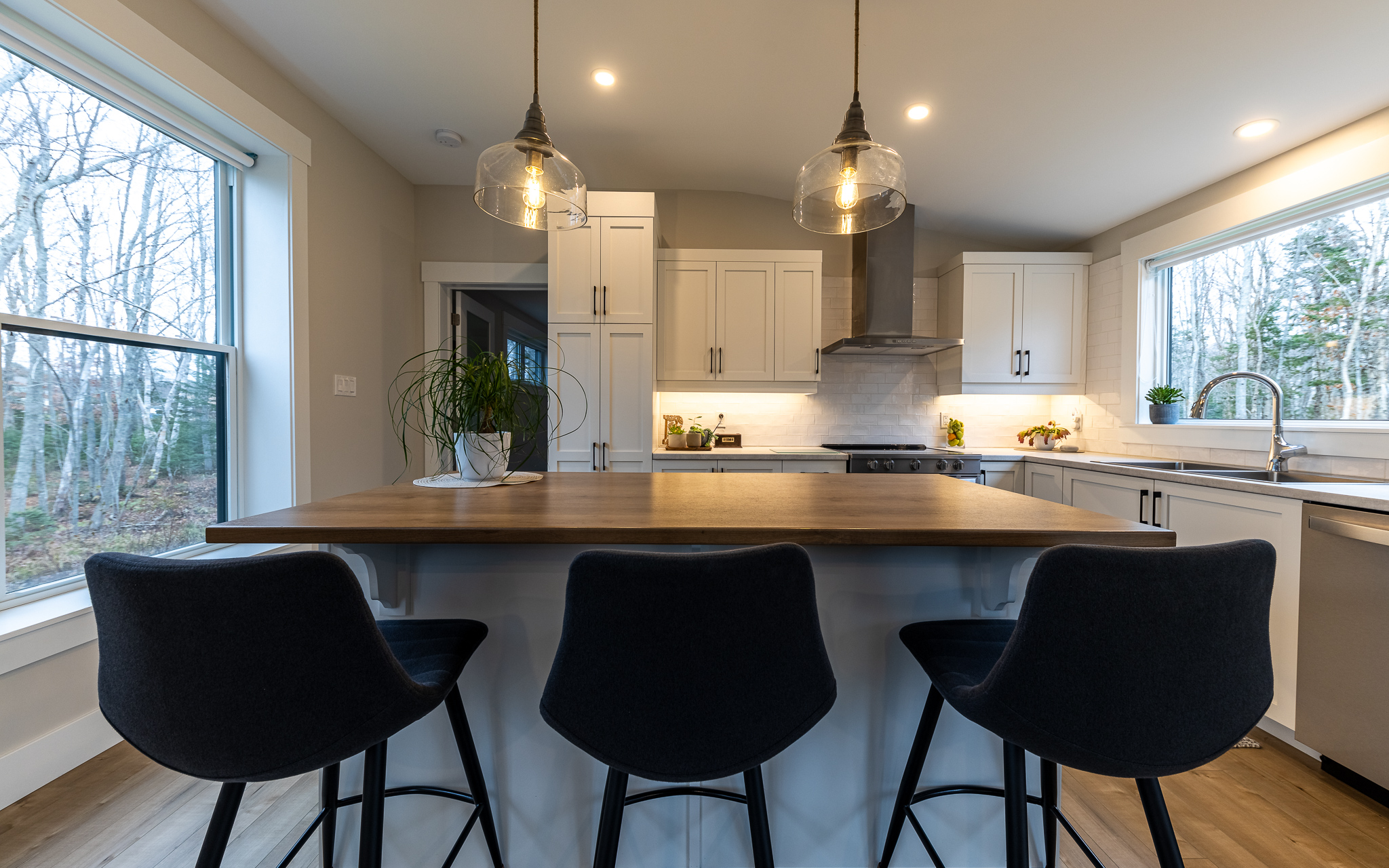Modern kitchen with a cozy ambiance, featuring a wooden island with three black chairs. Pendant lights hang above, and large windows offer a view of bare trees.
