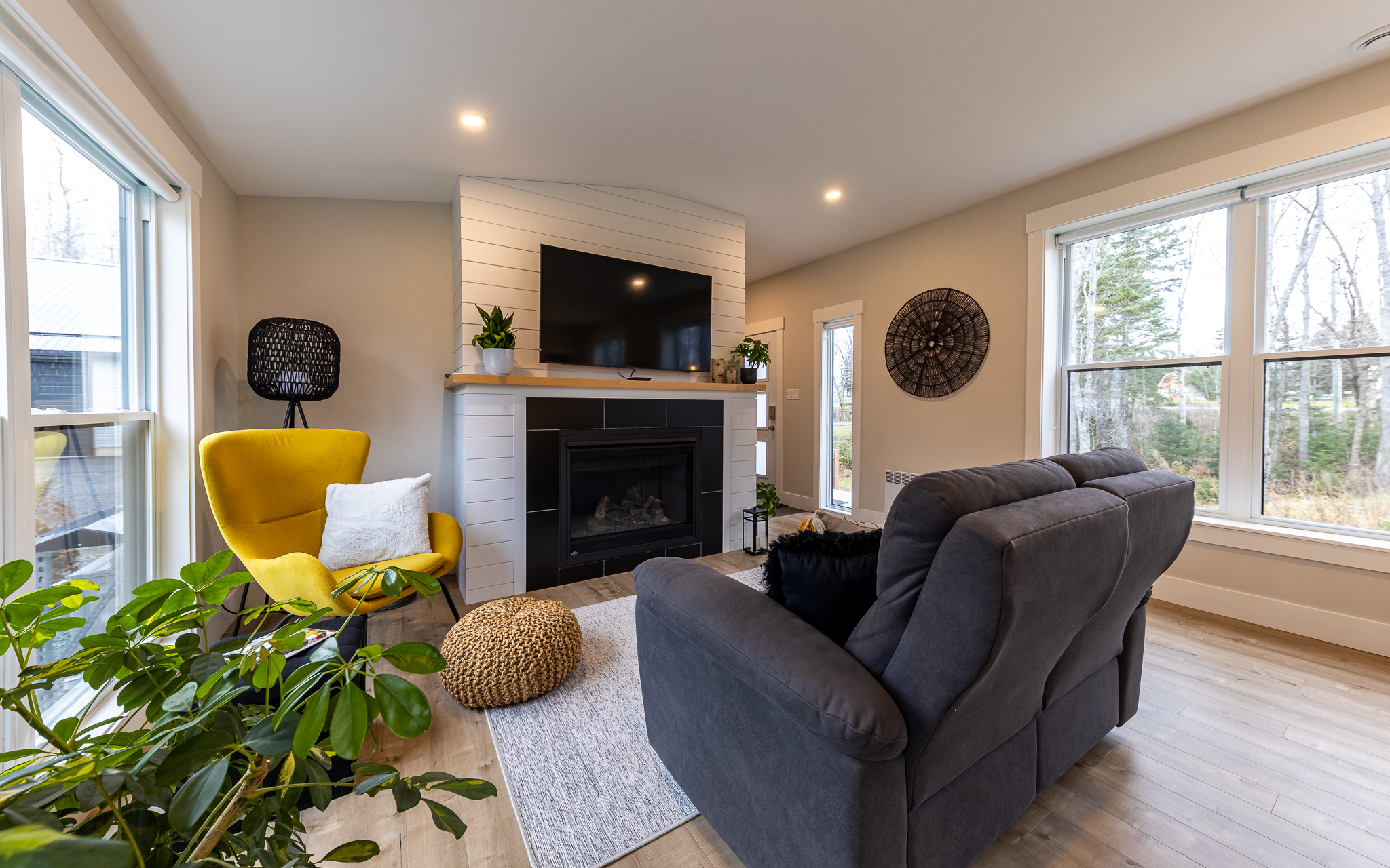 A cozy living room with a gray couch facing a fireplace and TV. A bright yellow chair adds a pop of color. Large windows provide natural light.
