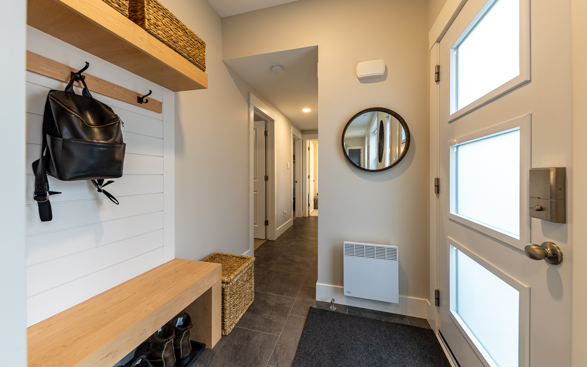 Modern entryway with a light wood bench, black backpack, round mirror, and woven baskets. The area conveys a neat, welcoming, and organized tone.