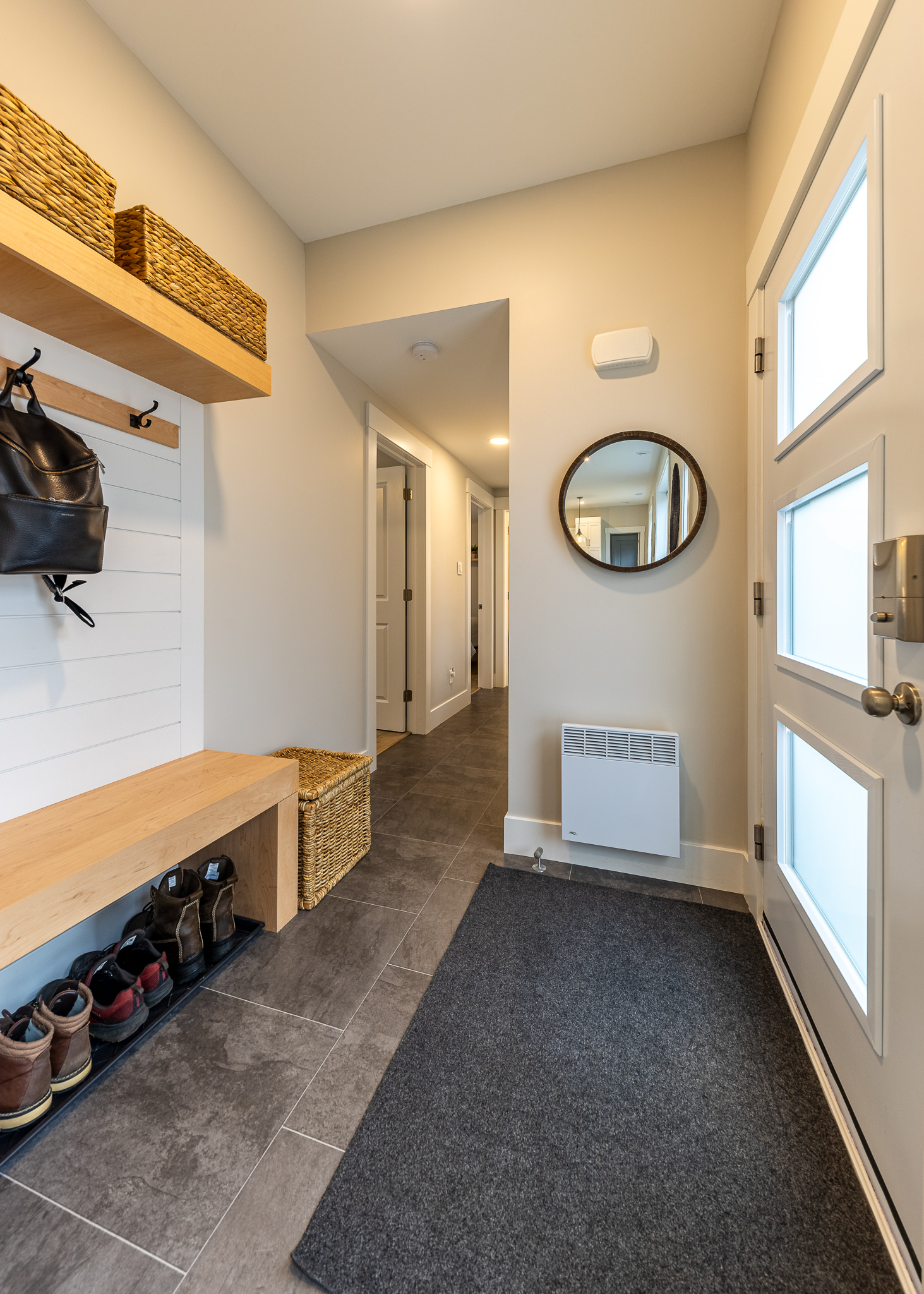 Compact, modern entryway with a wooden bench, shoe rack, and wicker baskets. A round mirror reflects light, adding warmth to neutral walls and gray tile floor.
