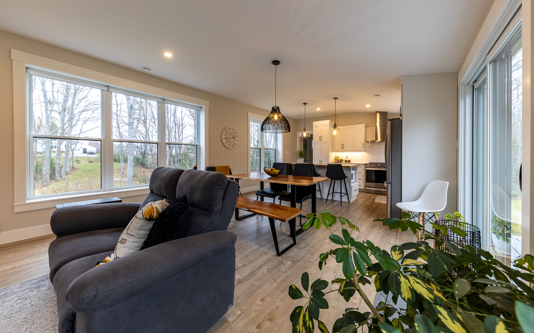 A cozy open-plan living area features a comfy gray armchair, wooden dining table, modern kitchen, and large windows with scenic outdoor views.