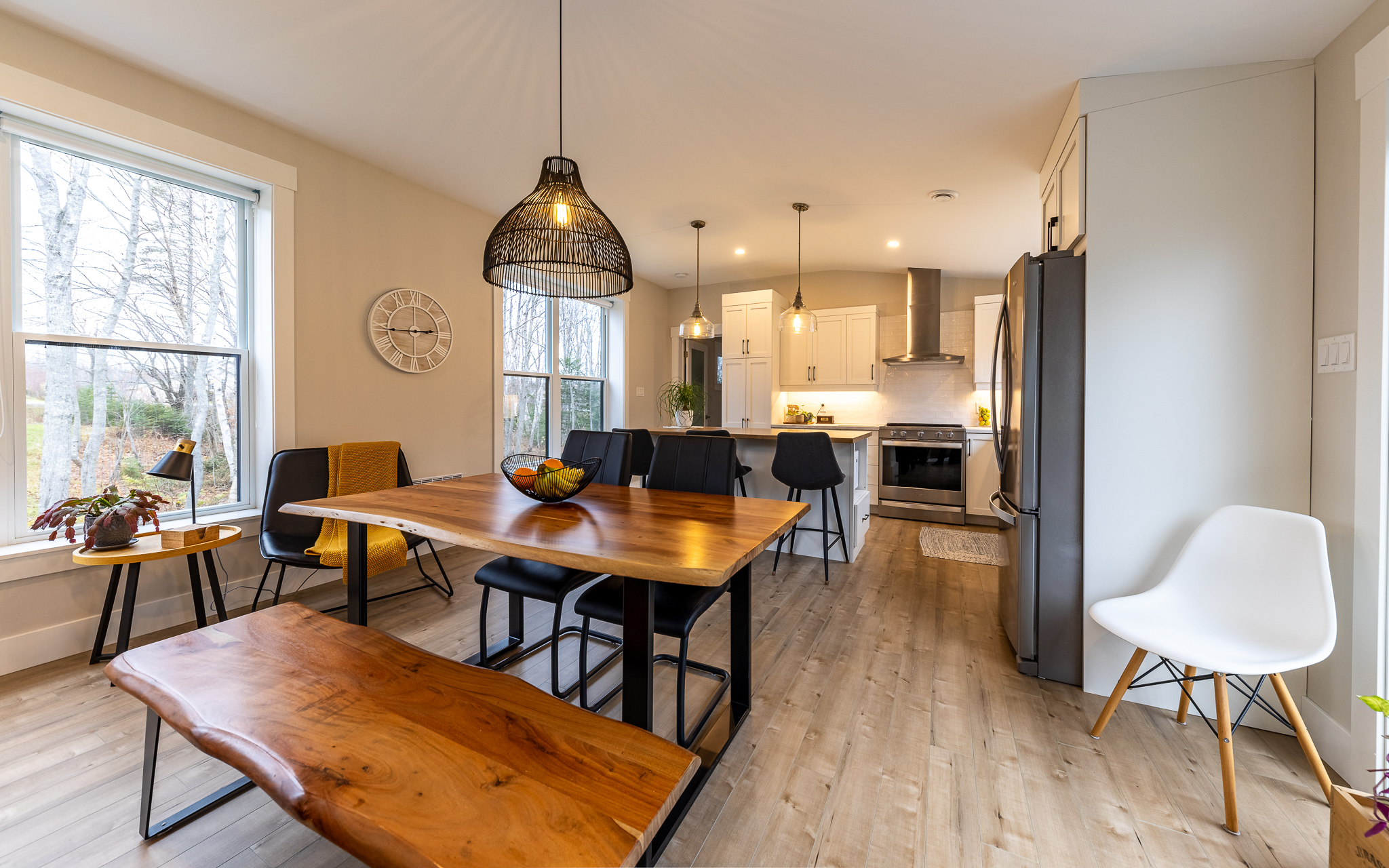 Modern dining room and kitchen with a wooden table and bench, black chairs, pendant lighting, and stainless steel appliances. Bright, inviting ambiance.