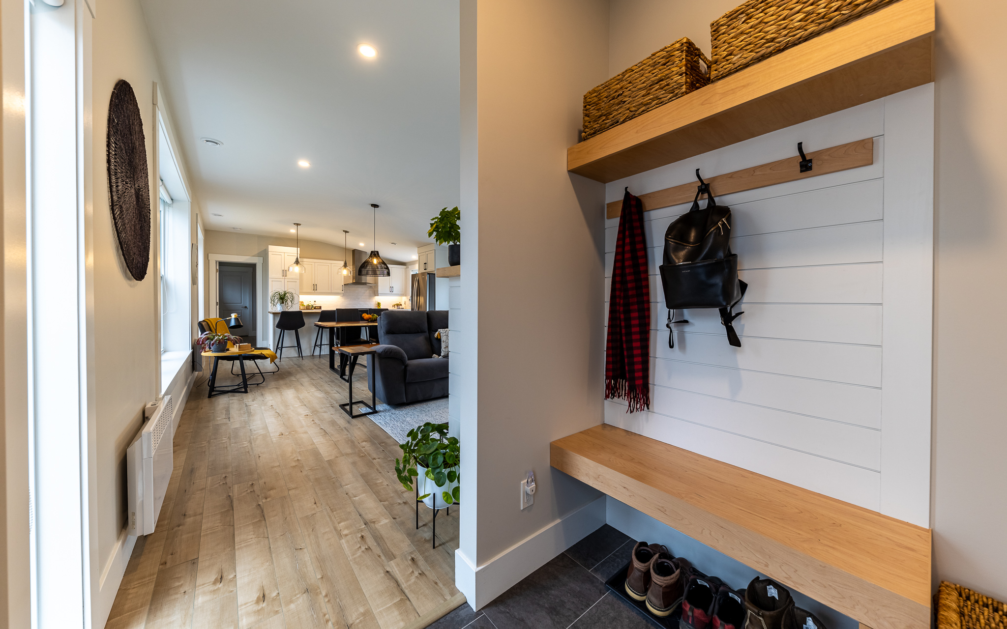 Modern entryway leading to a cozy, open-plan living room and kitchen. Wooden floors, pendant lights, a bench with hooks, and warm, inviting tones.