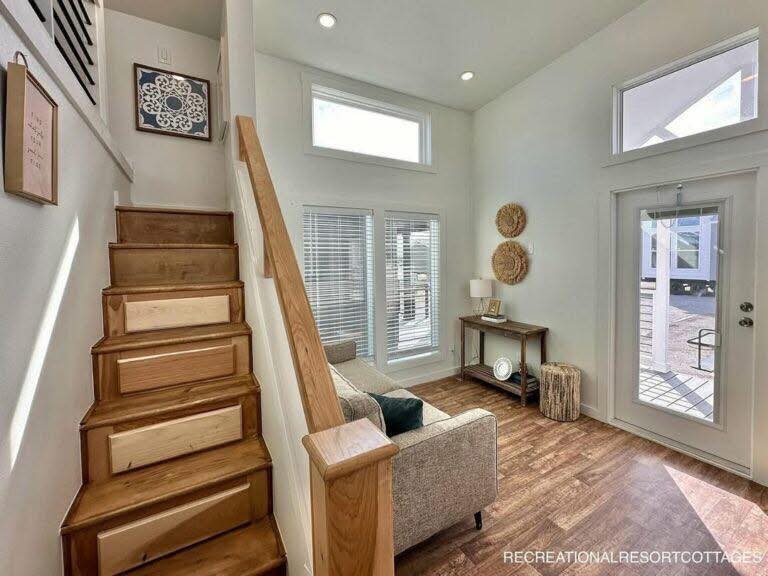 Modern tiny home interior with wooden stairs, natural light from tall windows, a cozy gray sofa, and a simple console table, creating an airy, inviting atmosphere.