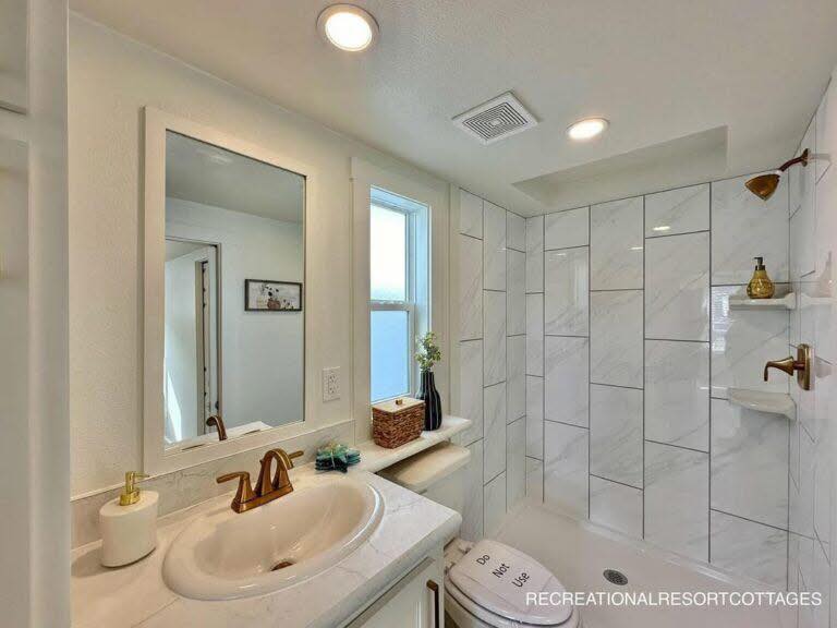 Modern bathroom with white tiled shower, brass fixtures, and a sink. A large mirror and window provide brightness. Calm, clean, and minimalist design.