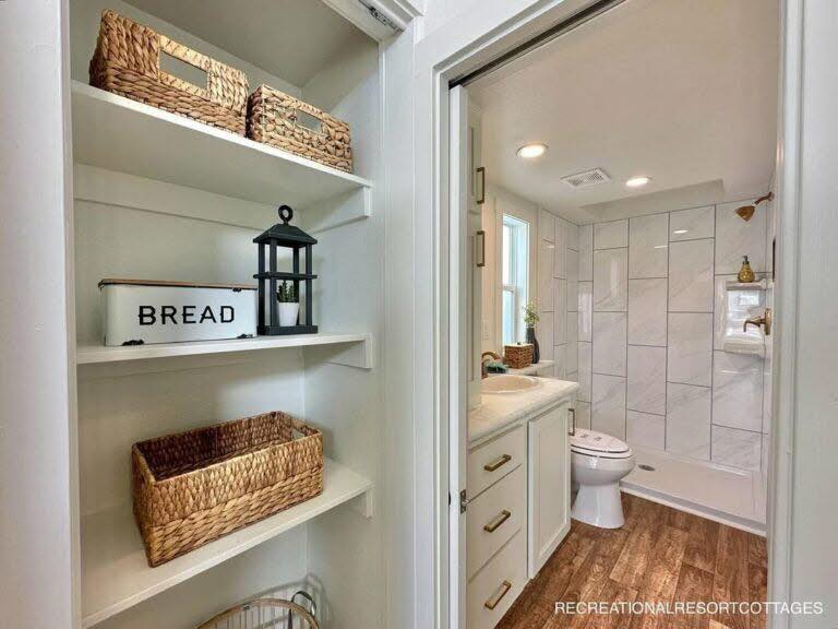 Shelves with woven baskets and decor lead to a bathroom with wooden floors, white cabinetry, a toilet, and a tiled shower, conveying a cozy, rustic feel.