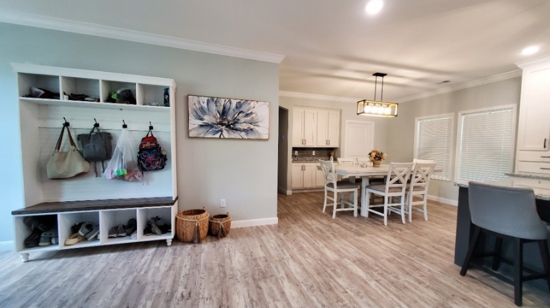 A modern dining area with light wood floors and neutral tones. On the left, a coat rack with bags, shoes, and a floral artwork above. A dining table with chairs is in the center under a rectangular light fixture. Wicker baskets and large windows add warmth.
