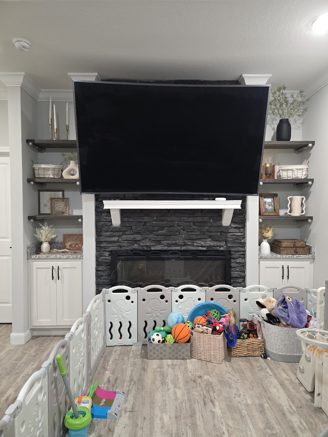 A spacious living room features a large, wall-mounted TV above a stone fireplace. Shelves with decor flank the fireplace. A play area with toys and a safety gate occupies the foreground, reflecting a cozy and family-friendly atmosphere.