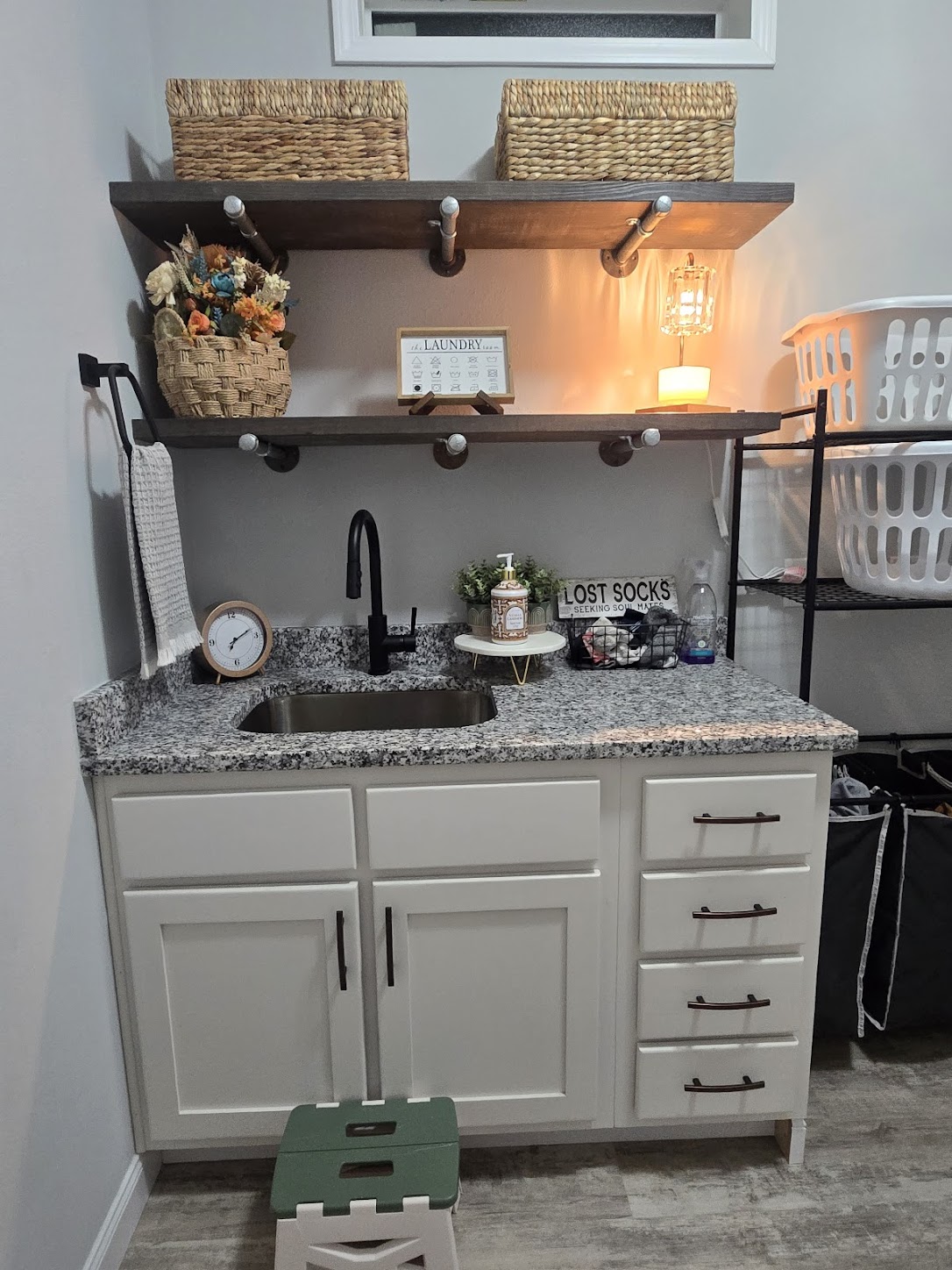 Laundry room with gray granite countertop, black faucet, and white cabinetry. Two wooden shelves hold baskets, a "Lost Socks" sign, and decor. Warm lighting adds a cozy feel.
