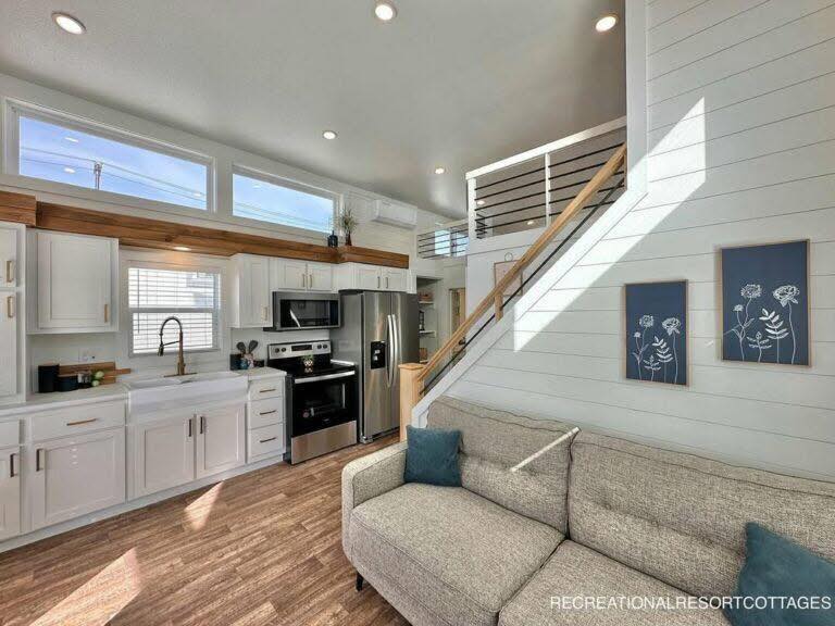 Modern tiny home interior with white kitchen cabinets, stainless steel appliances, wooden accents, and bright natural light. Cozy sofa near staircase.