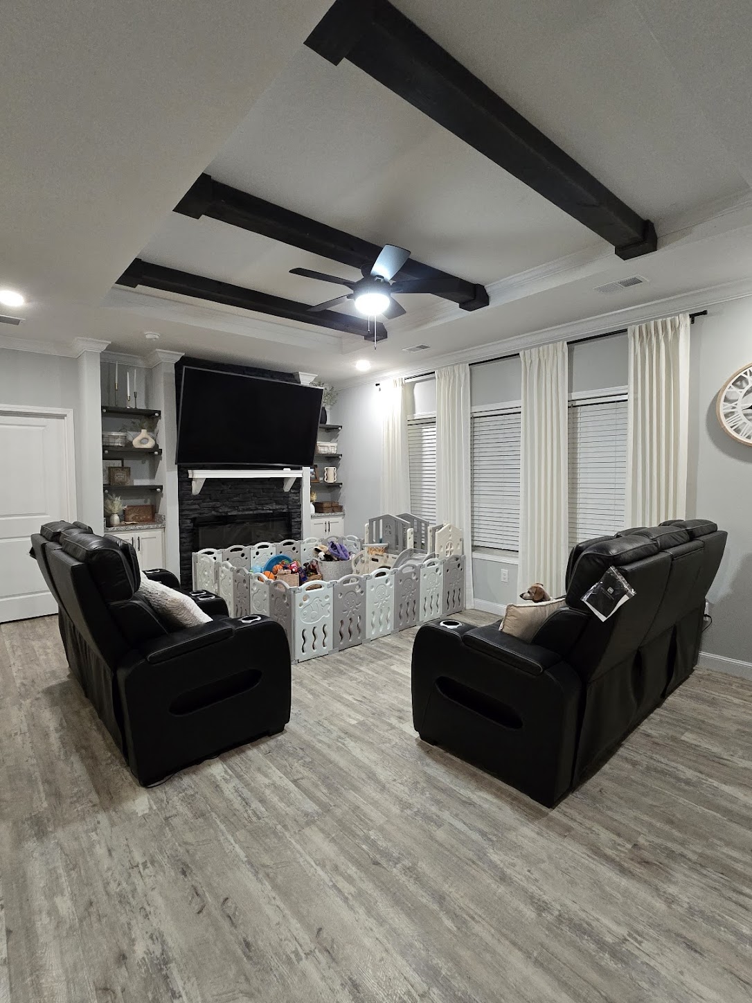 Stylish living room with dark beams, gray wood flooring, and a centered black sectional. A playpen filled with toys sits below a large TV. Bright and cozy.