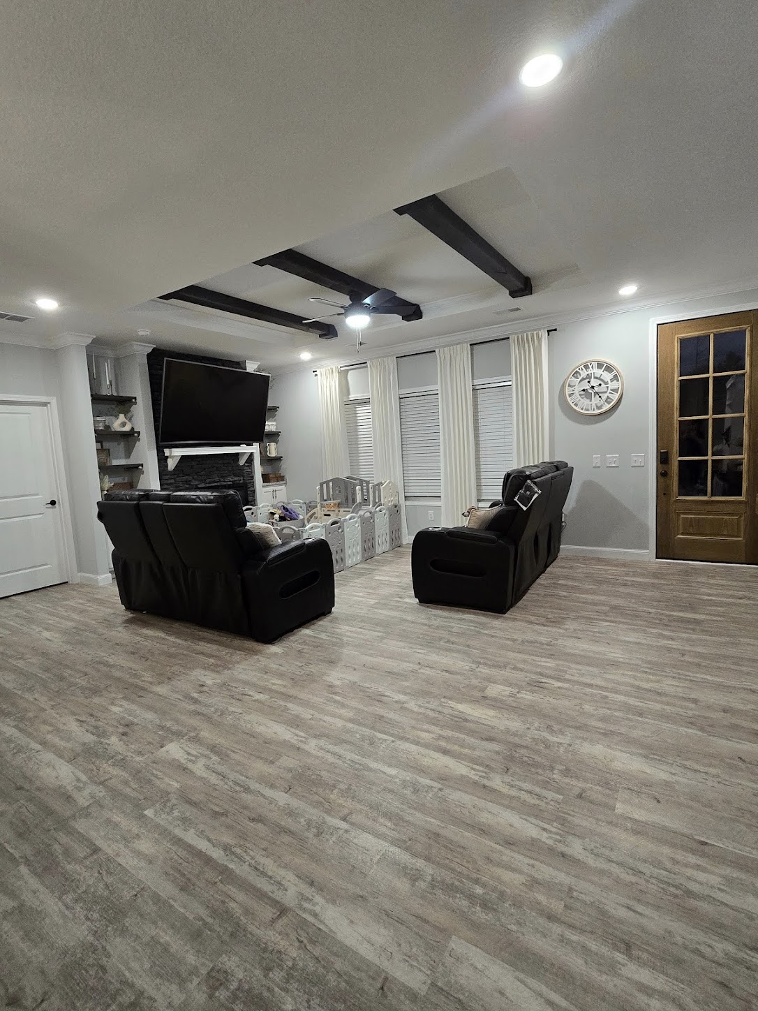 A cozy living room with dark sofas facing a wall-mounted TV, wooden floors, exposed ceiling beams, and a decorative wall clock. Comfortable and inviting.