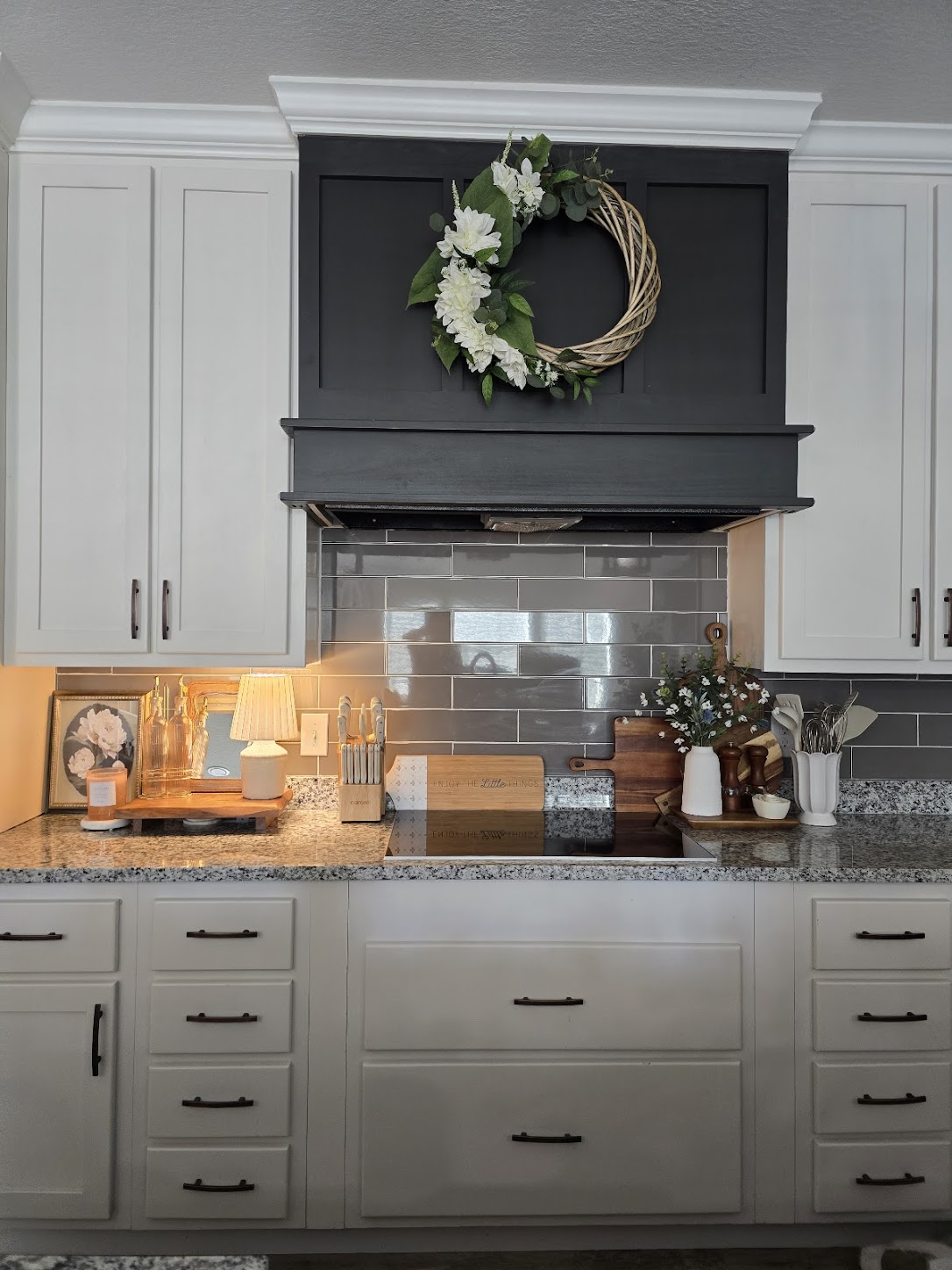 A cozy kitchen with white cabinets and a dark wood hood, featuring a wreath of white flowers and greenery. Granite countertops hold decor and utensils, creating a warm, inviting ambiance.