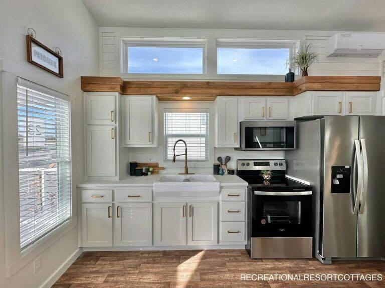 Modern kitchen with white cabinets, stainless steel appliances, farmhouse sink, and rustic wood accents under upper windows. Bright, airy ambiance.