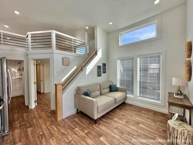 Bright, modern living room with wood flooring, a cozy gray sofa, and blue cushions. Large windows let in natural light, and a lofted space is visible above.