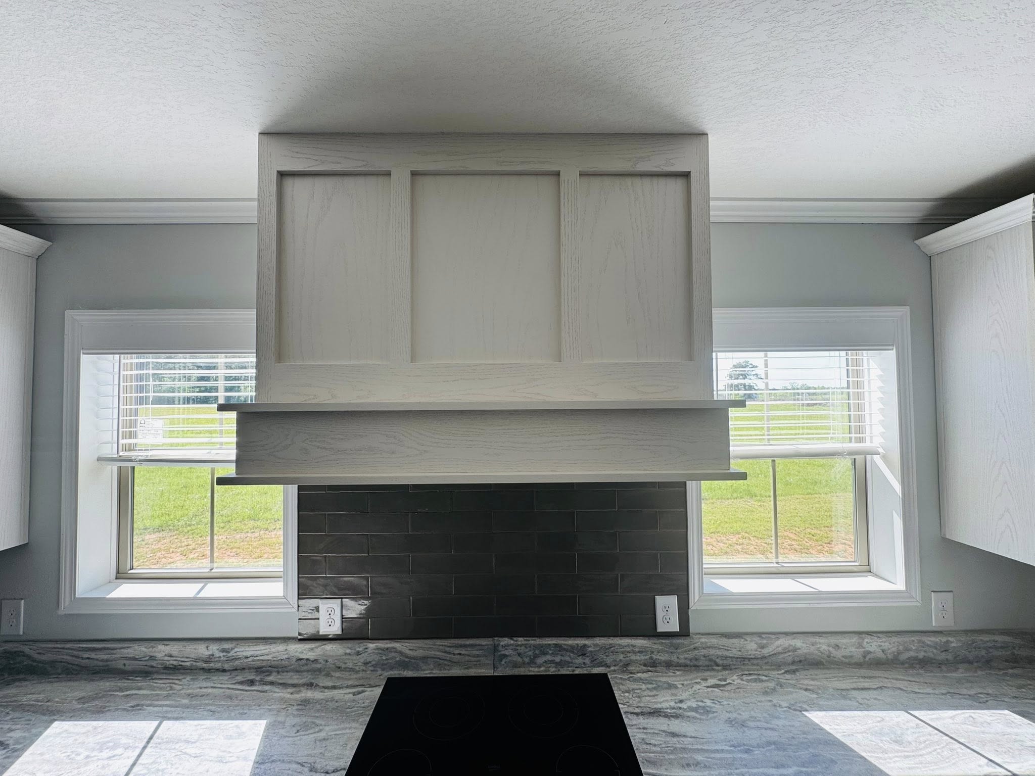 Bright kitchen with white cabinetry and a sleek black tile backsplash. Two windows frame the scene, offering a view of green grass outside.