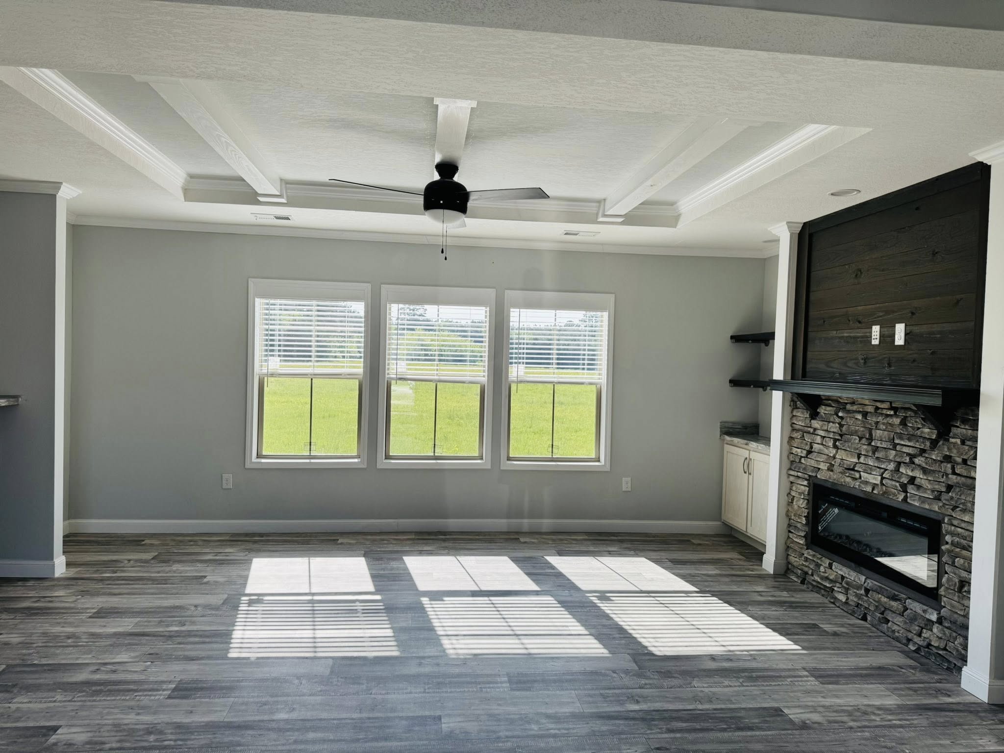 Spacious living room with gray walls and wood floor, featuring a large window with a sunny view, modern ceiling fan, and stone fireplace. Cozy and bright atmosphere.