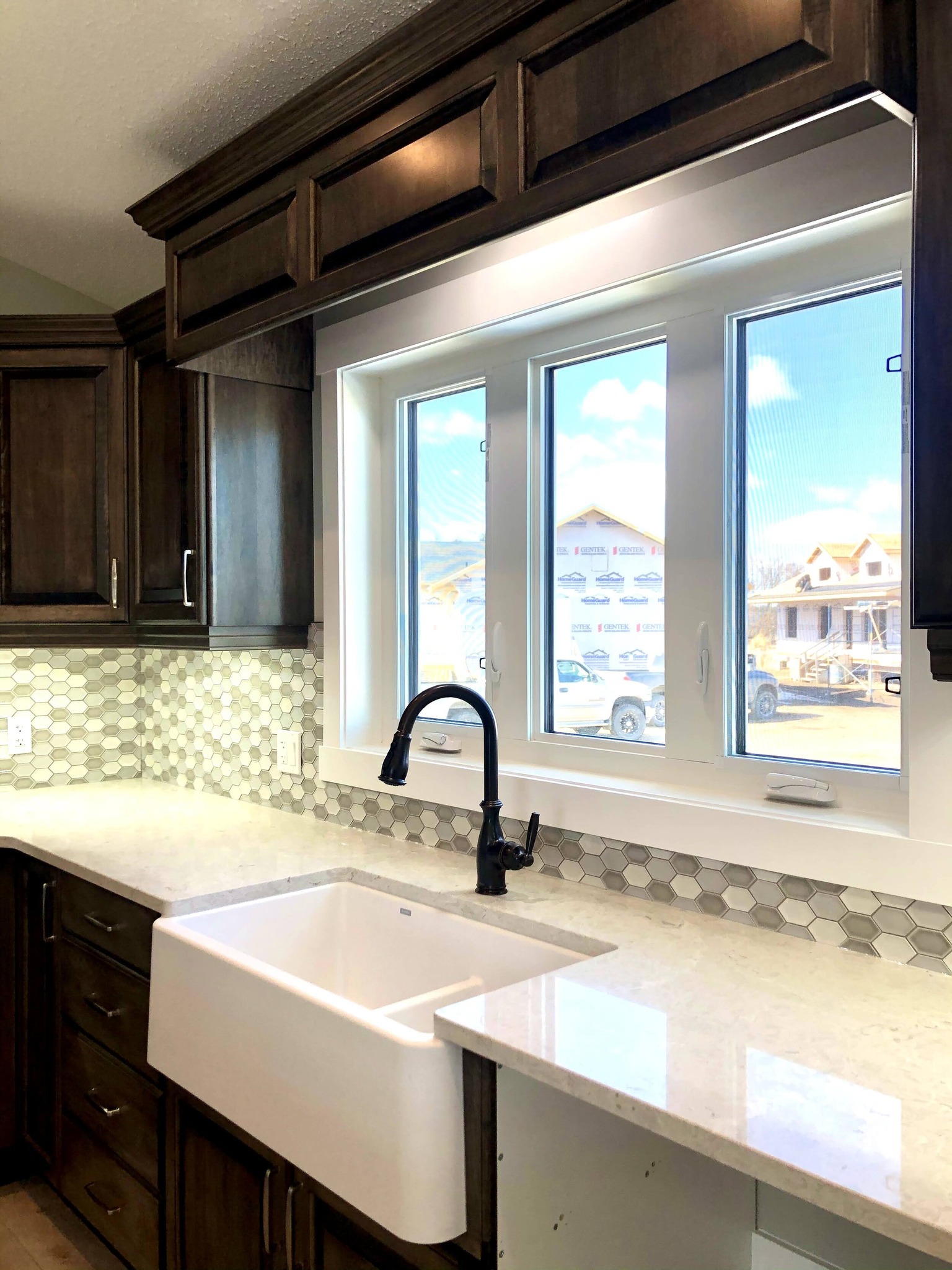 A bright kitchen with dark wood cabinets, hexagonal tile backsplash, and a white farmhouse sink. A window offers a view of a sunny, suburban scene.