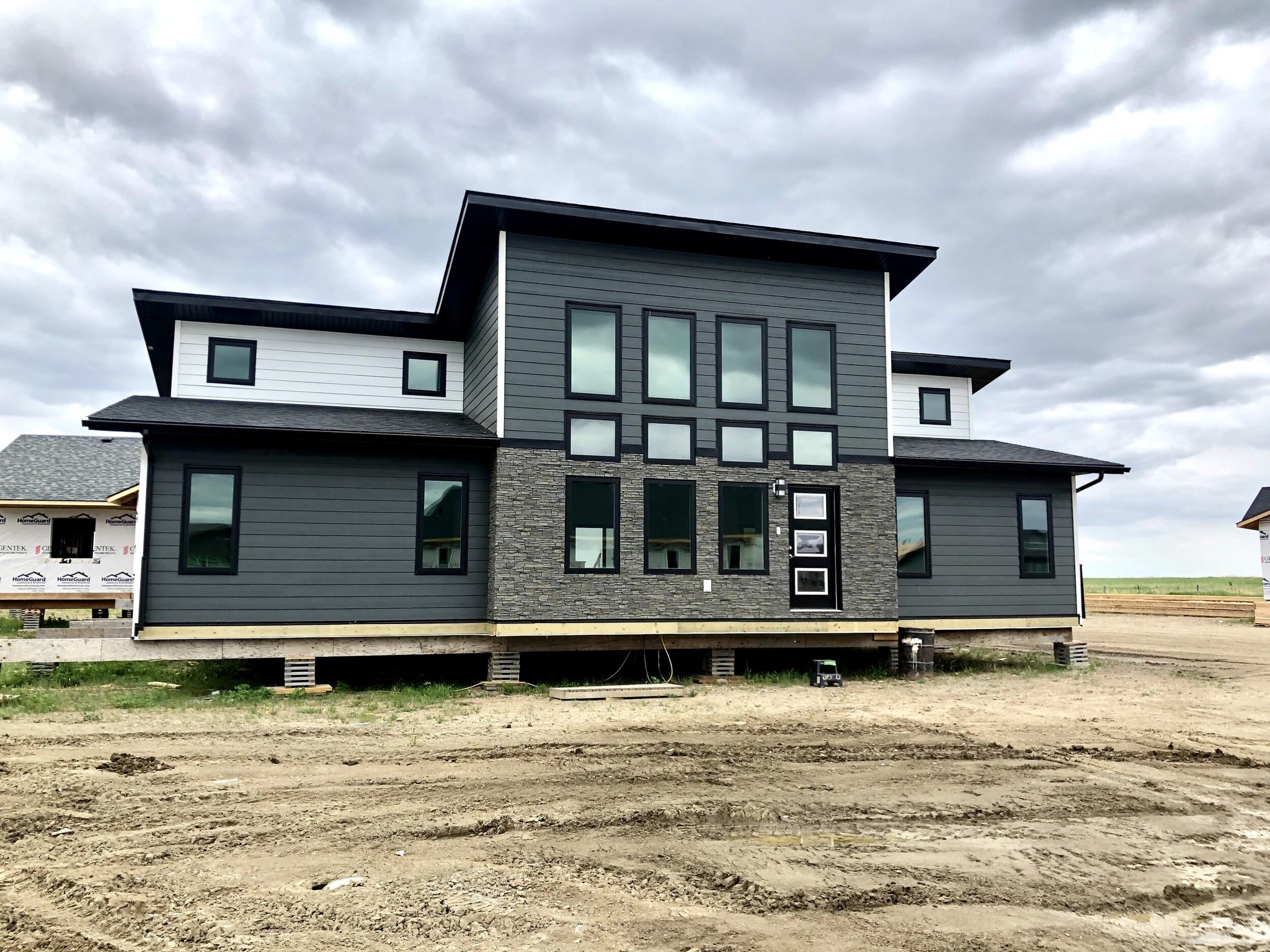Modern gray house under construction, featuring large windows and a flat roof. The sky is overcast, and the surrounding area is muddy and undeveloped.