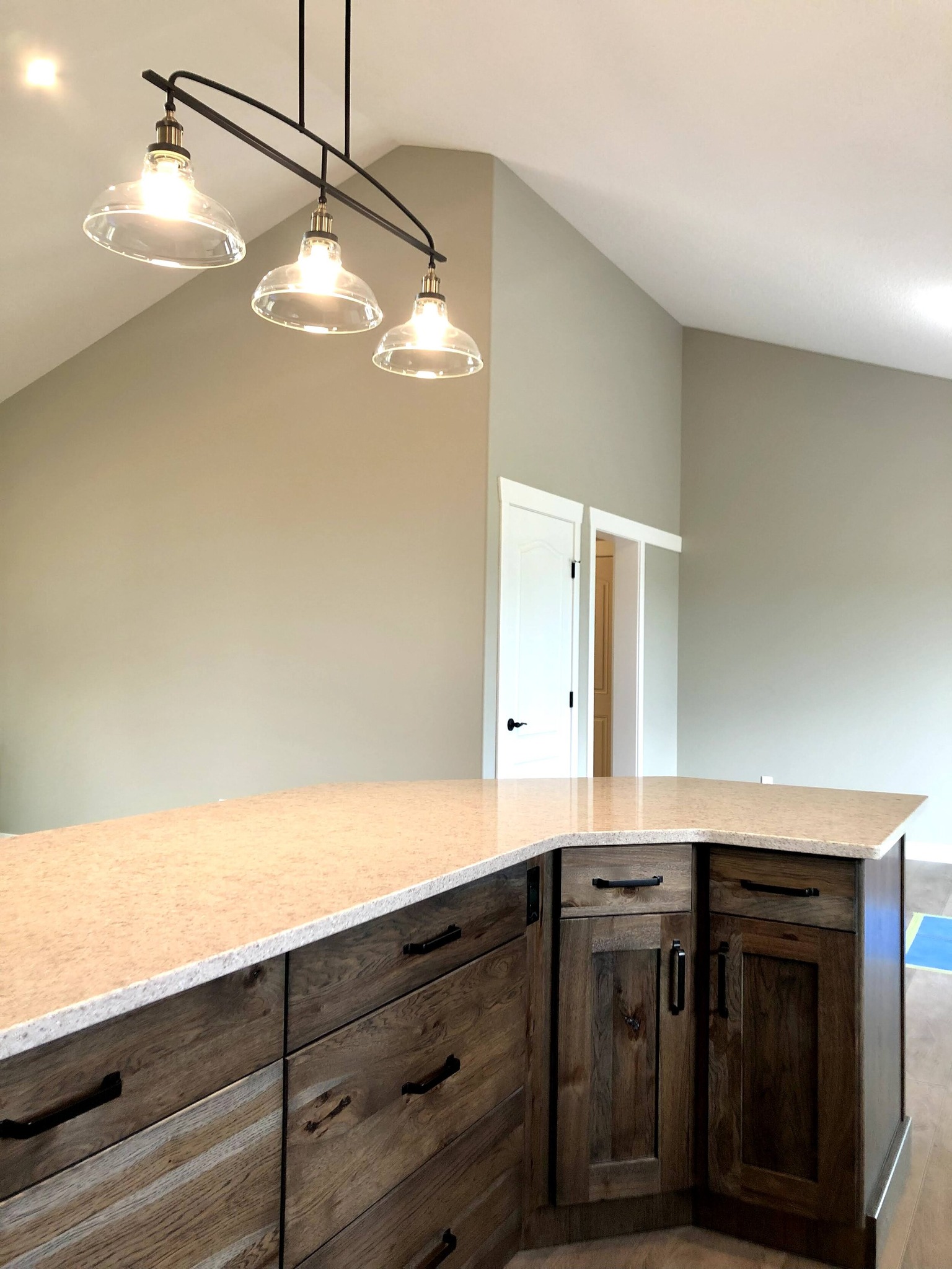 Kitchen with vaulted ceiling, featuring a light fixture with three bulbs above a light granite countertop and rustic wooden cabinetry. Cozy and modern.