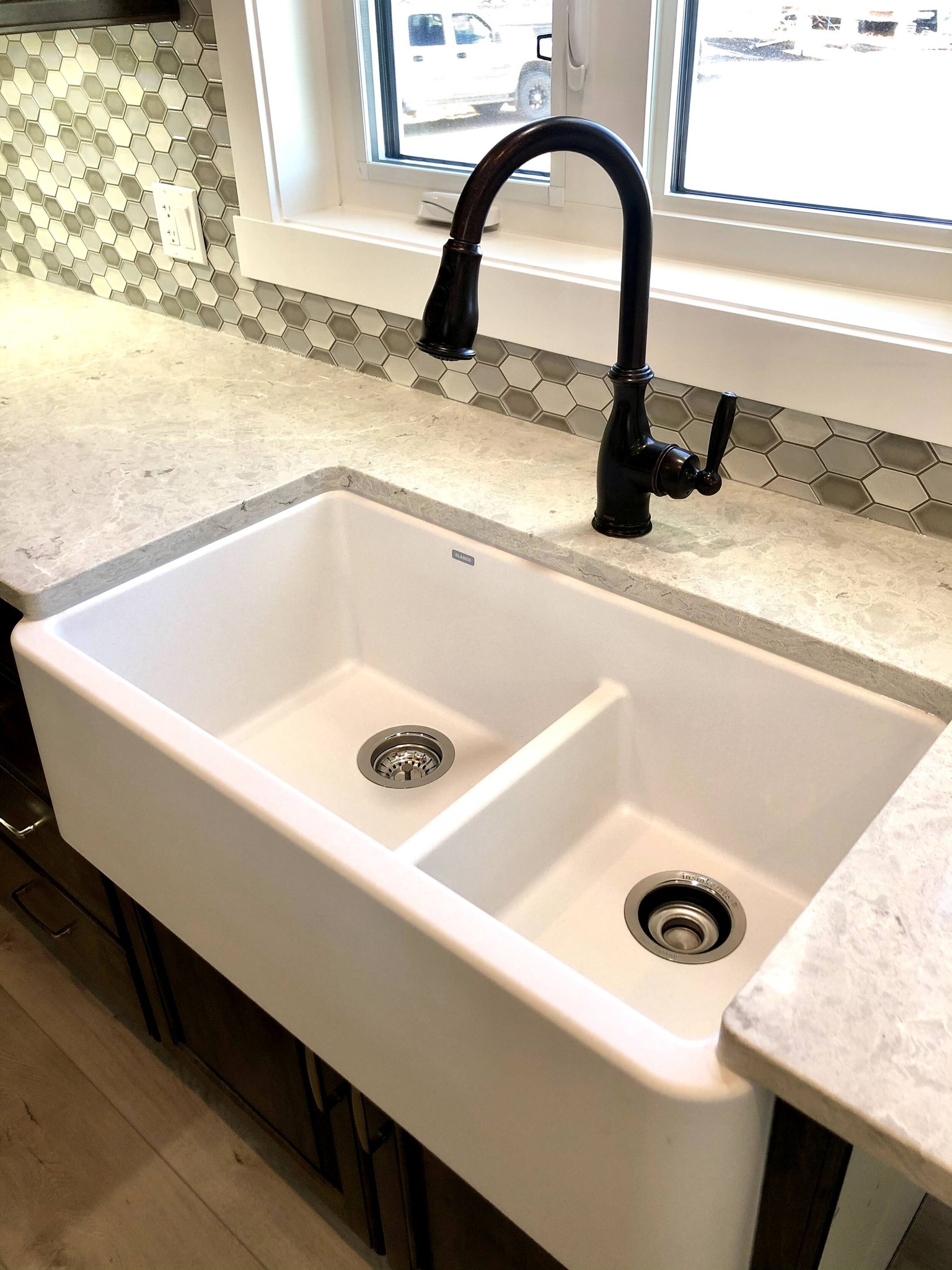 A farmhouse-style white double sink with a black faucet sits under a window. The countertop is light marble, and the backsplash features hexagonal tiles.