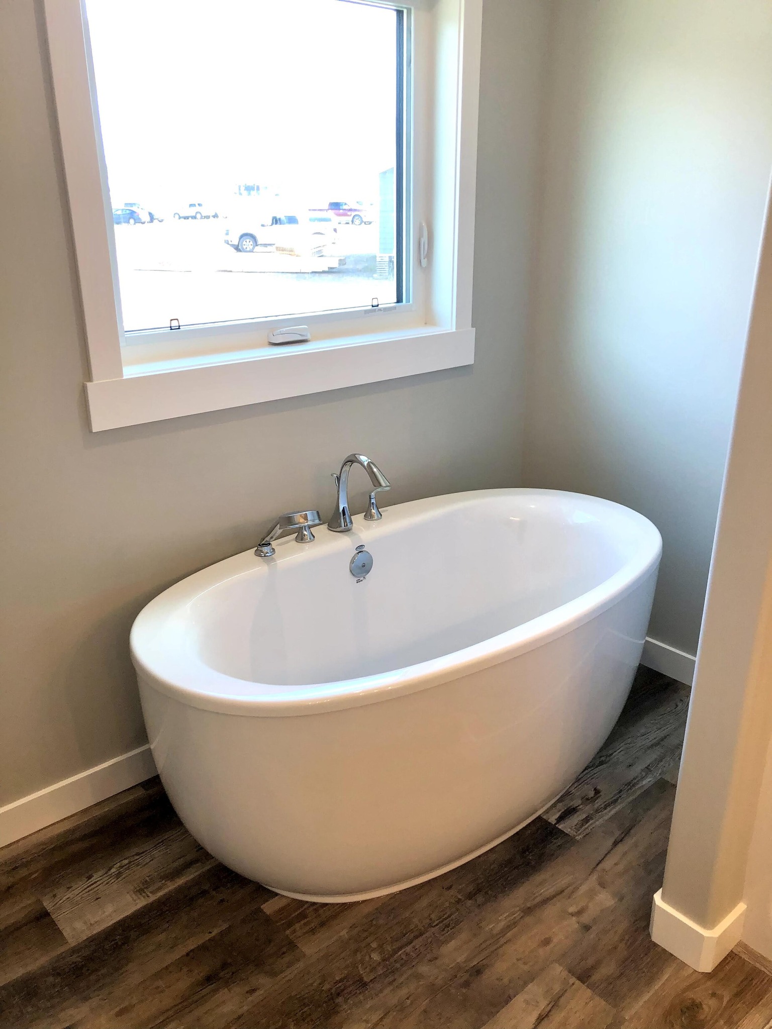Freestanding white bathtub with chrome fixtures by a window in a bright bathroom. Light wood floor and neutral walls create a calm ambiance.