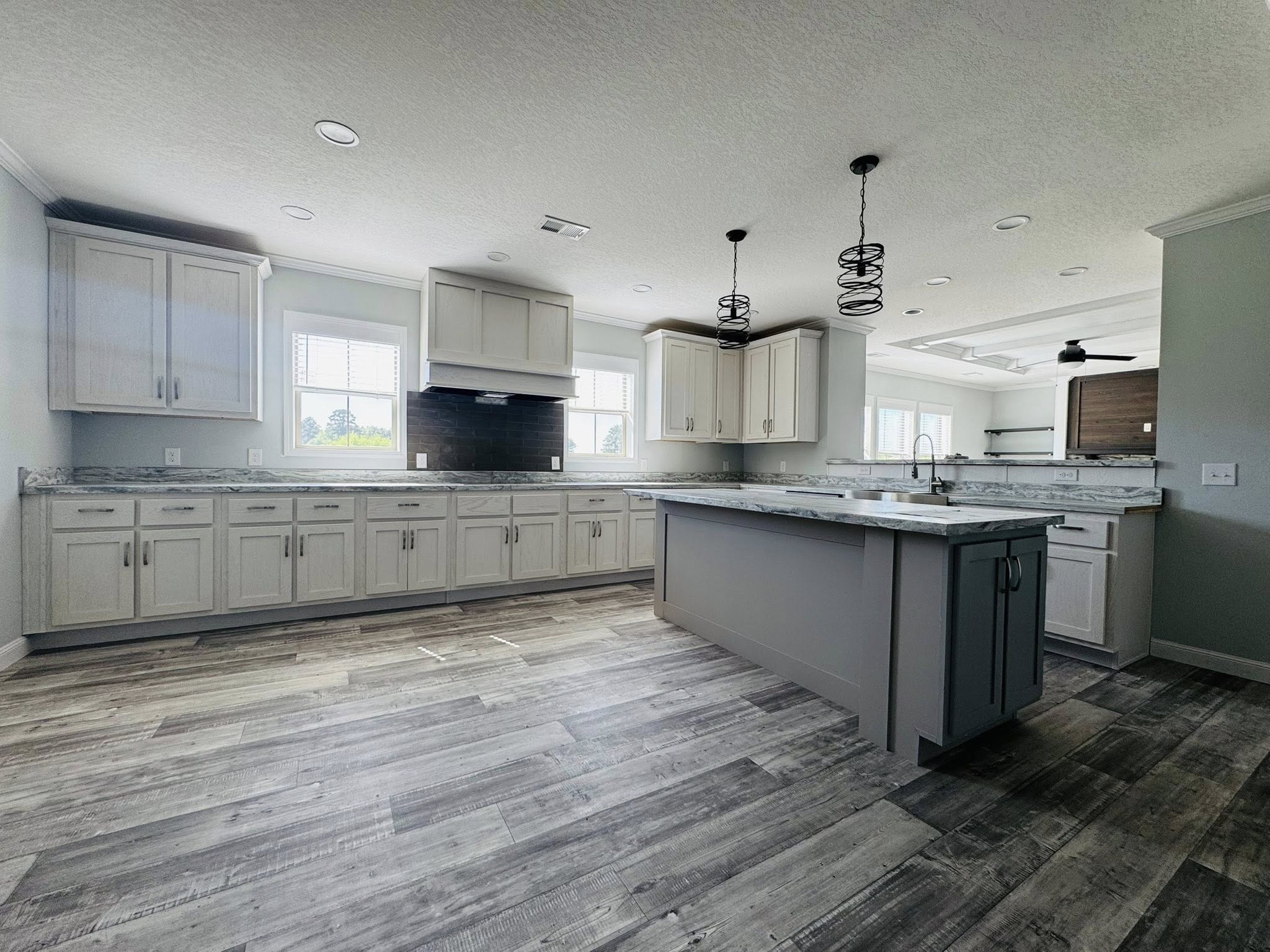 Spacious, modern kitchen with gray cabinets, marble countertops, and pendant lights. Bright windows, wood floor, island, and sleek ambiance.