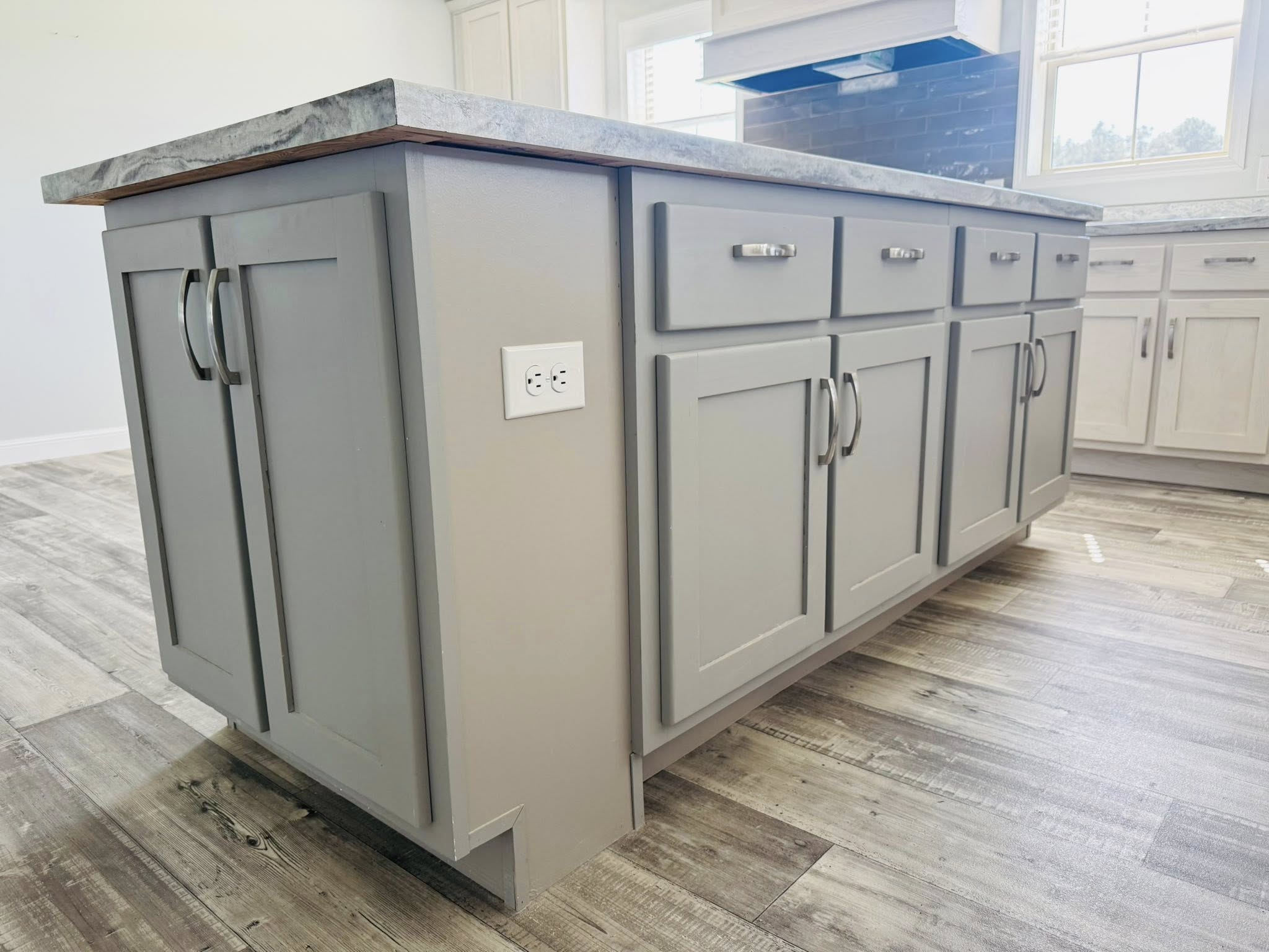 A modern kitchen island with gray paneled cabinets, silver handles, and a marble countertop. Light wood flooring and a power outlet are visible.
