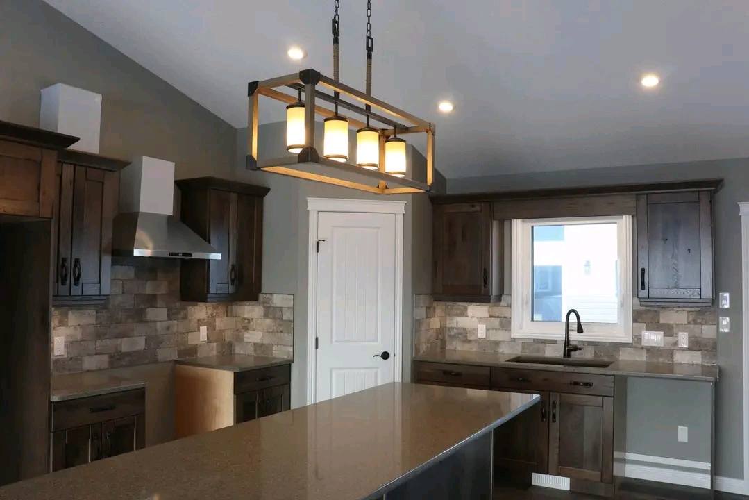 Modern kitchen with dark wood cabinets, gray stone backsplash, and central island. A rectangular pendant light hangs overhead, creating a warm ambiance.
