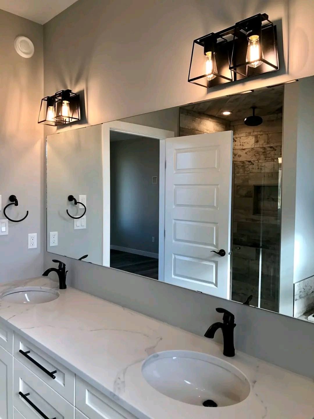 Modern bathroom with a double sink vanity. Black faucets and towel rings contrast with white surfaces. Warm lighting above mirrors creates a cozy feel.