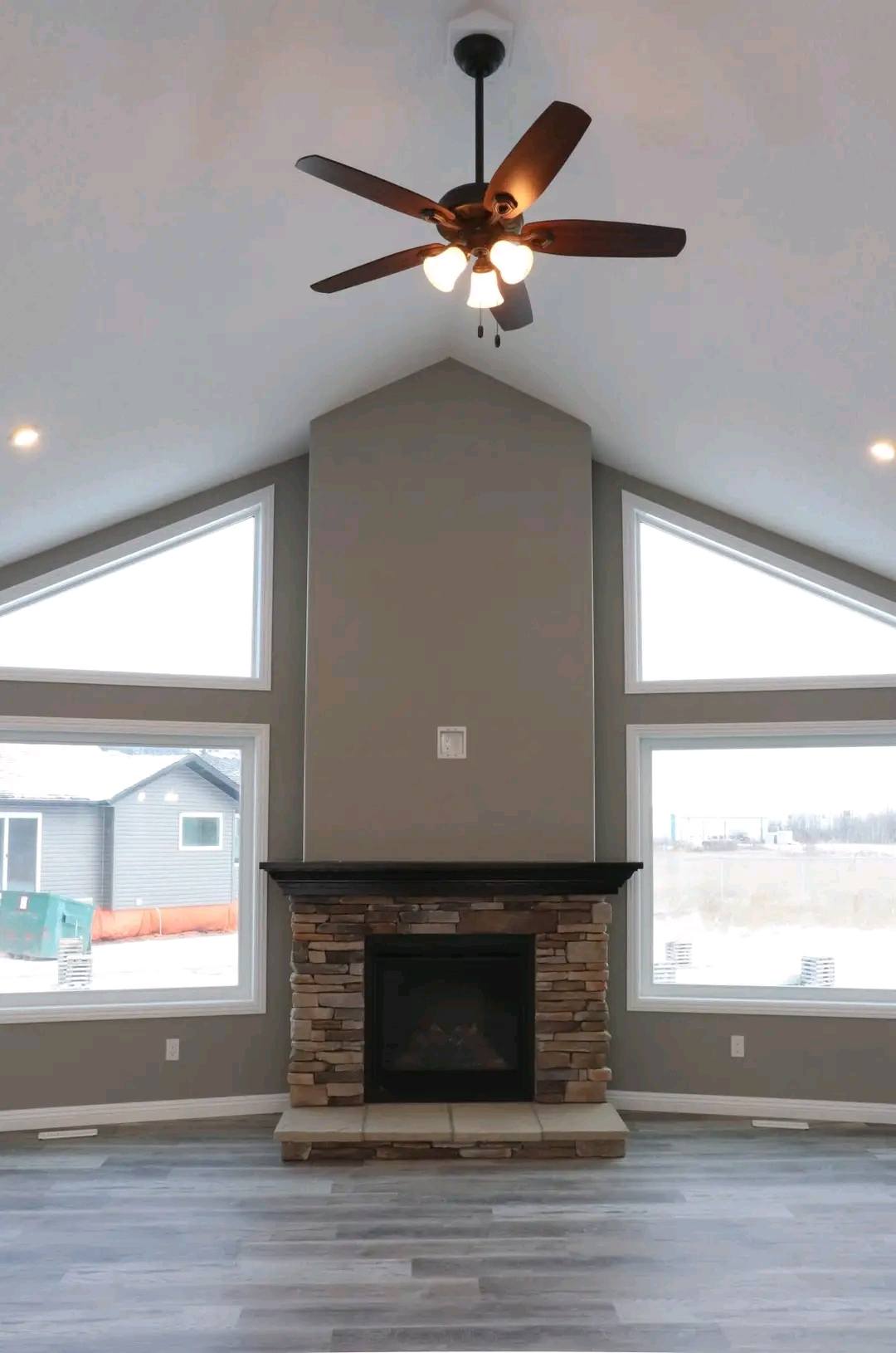 Spacious room with vaulted ceiling, gray walls, stone fireplace, modern ceiling fan, and large triangular windows. Bright and airy ambiance.