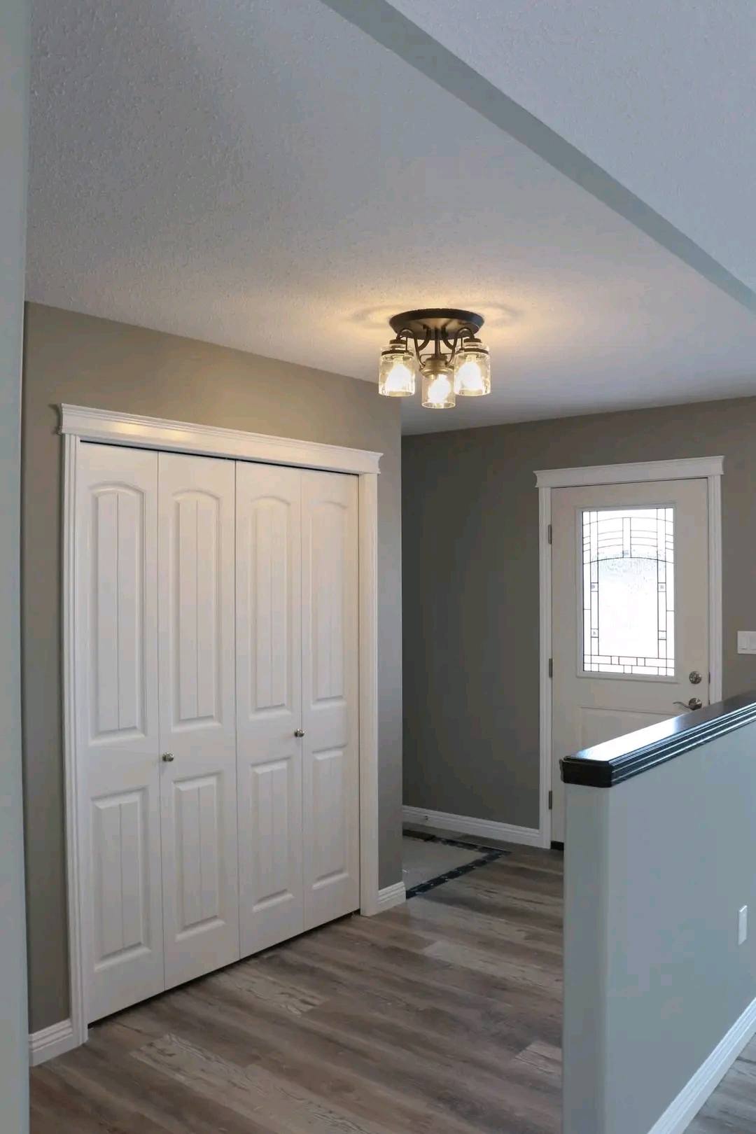 A hallway with a wooden floor, white double closet doors on the left, a white door with decorative glass on the right, and a ceiling light fixture.