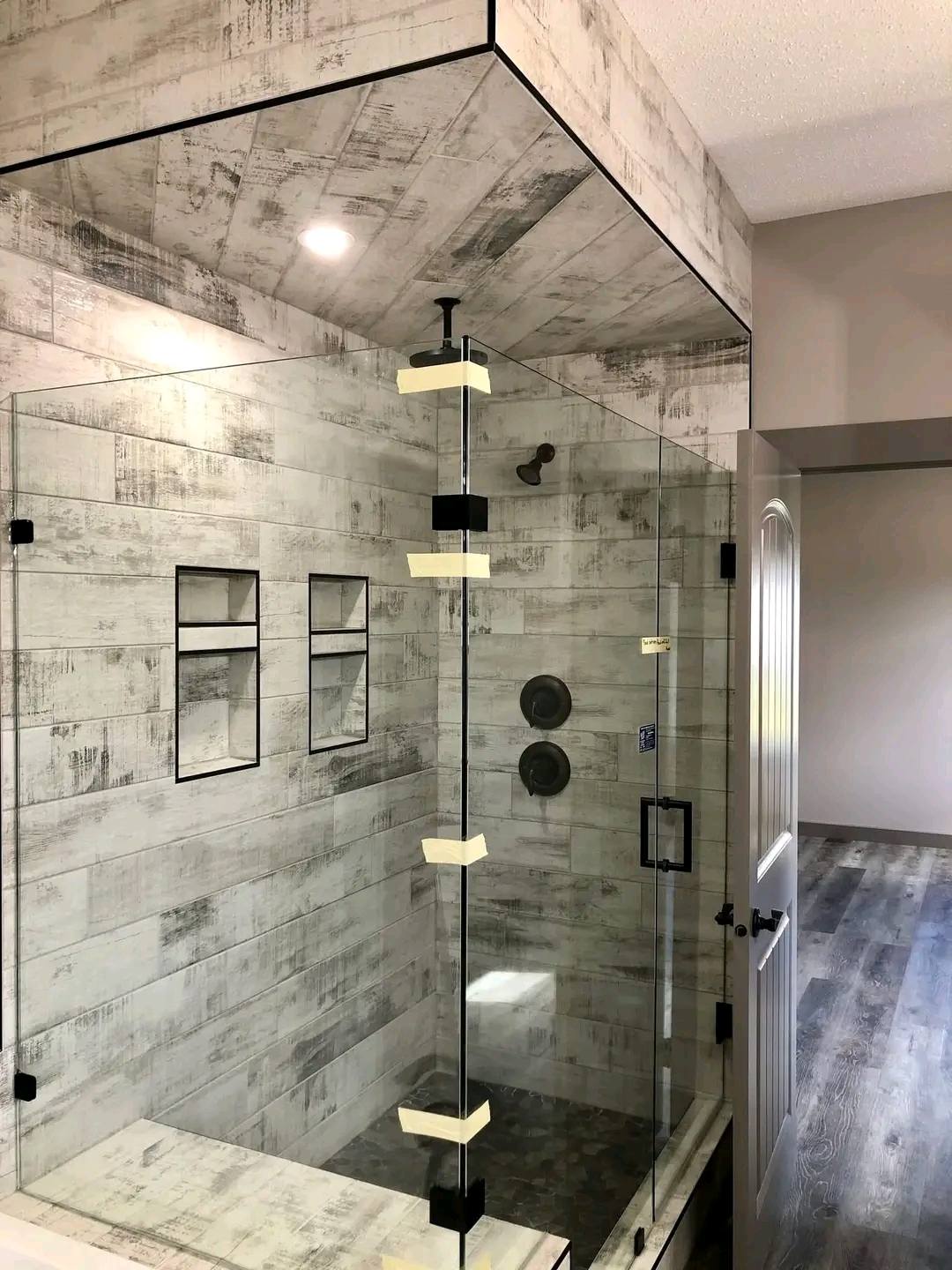 Modern shower with glass doors and a rustic wood-textured tile interior. Features built-in shelves, dual showerheads, and a serene, minimalist style.