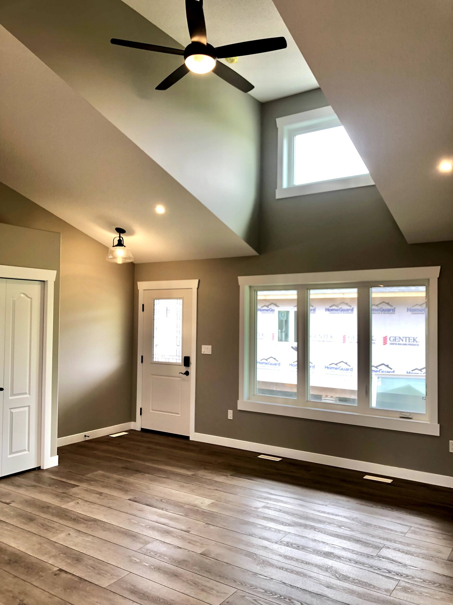 Spacious room with vaulted ceiling, ceiling fan, and modern light fixtures. Features hardwood floors, a white door, and a large window. Bright and open atmosphere.