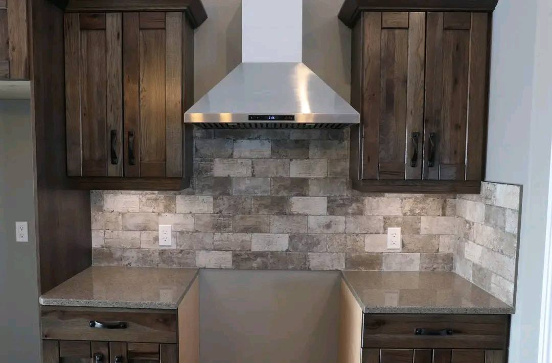 Modern kitchen with gray stone tile backsplash, stainless steel vent hood, and dark wood cabinets. Smooth countertops with warm, inviting lighting.
