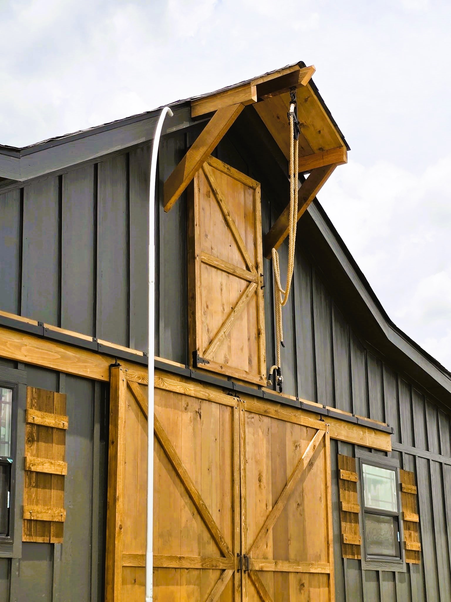 A modern barn with dark gray siding and wooden sliding doors featuring a rustic design. The structure includes small windows and a pulley system, under a cloudy sky.