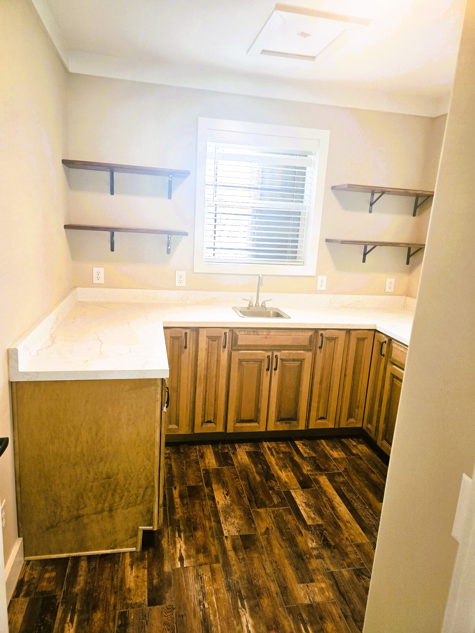 A small, modern kitchen with light wood cabinets and dark wooden floors. Marble countertops, a centered sink, and wall-mounted shelves create a tidy, warm atmosphere.