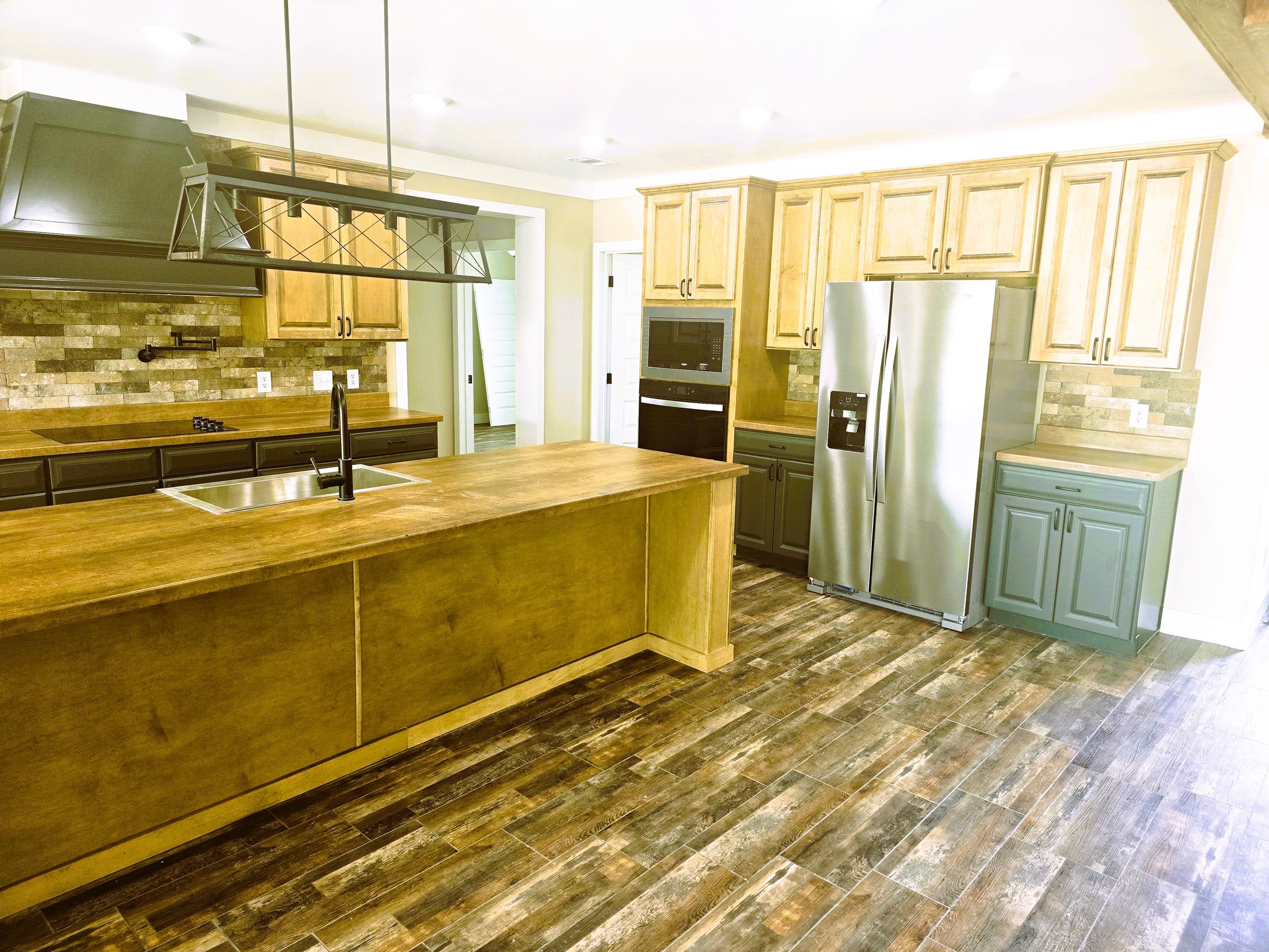 Spacious kitchen with wood cabinets and island, stainless steel fridge, modern light fixture, and stone-tiled backsplash. Warm, rustic atmosphere.