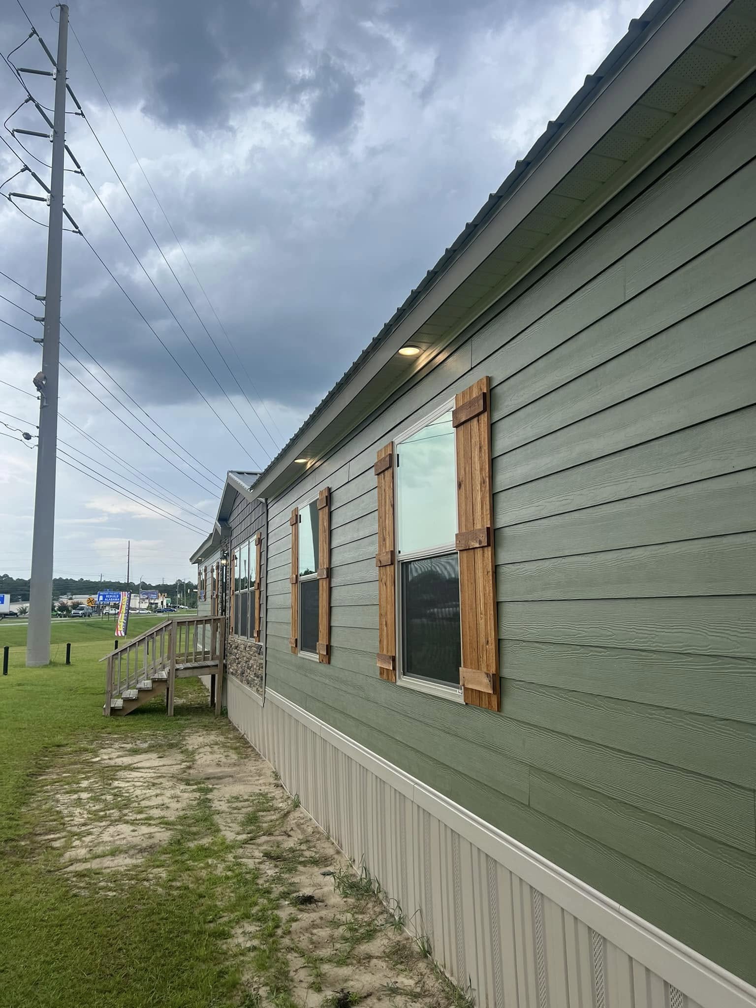 Side view of a green modular home with brown shutters and a stone accent wall. Overcast sky and power lines create a calm, pre-storm atmosphere.