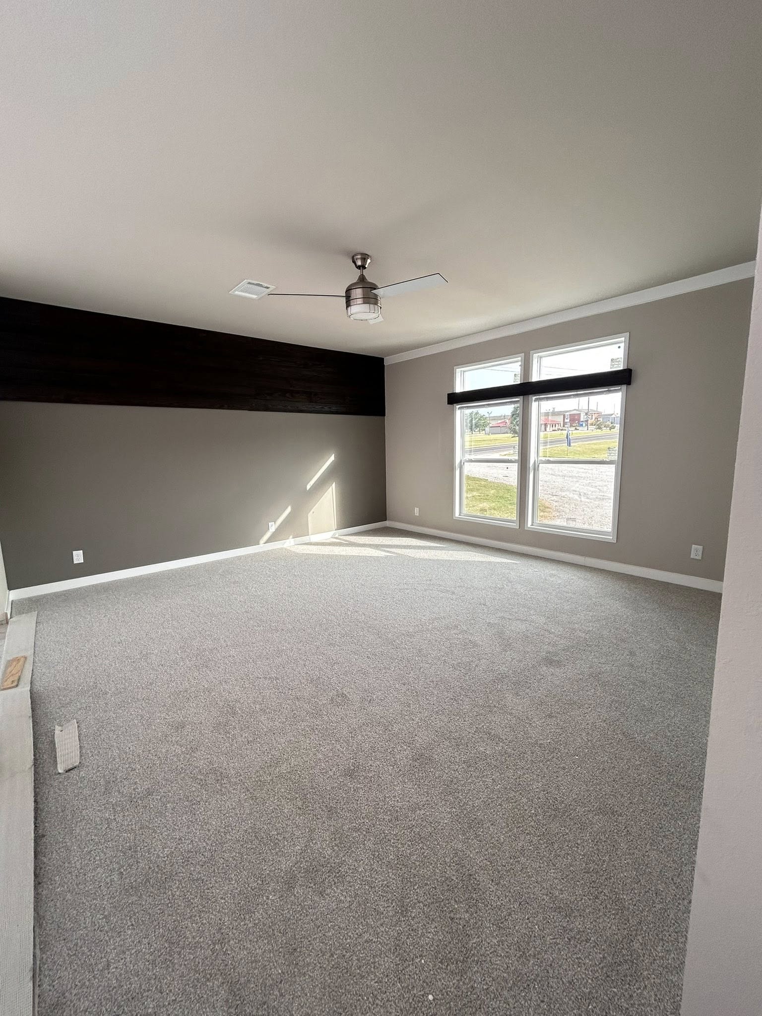 Empty room with gray carpet, a dark accent wall, and large windows letting in natural light. Ceiling fan in the center, creating a calm, spacious feel.