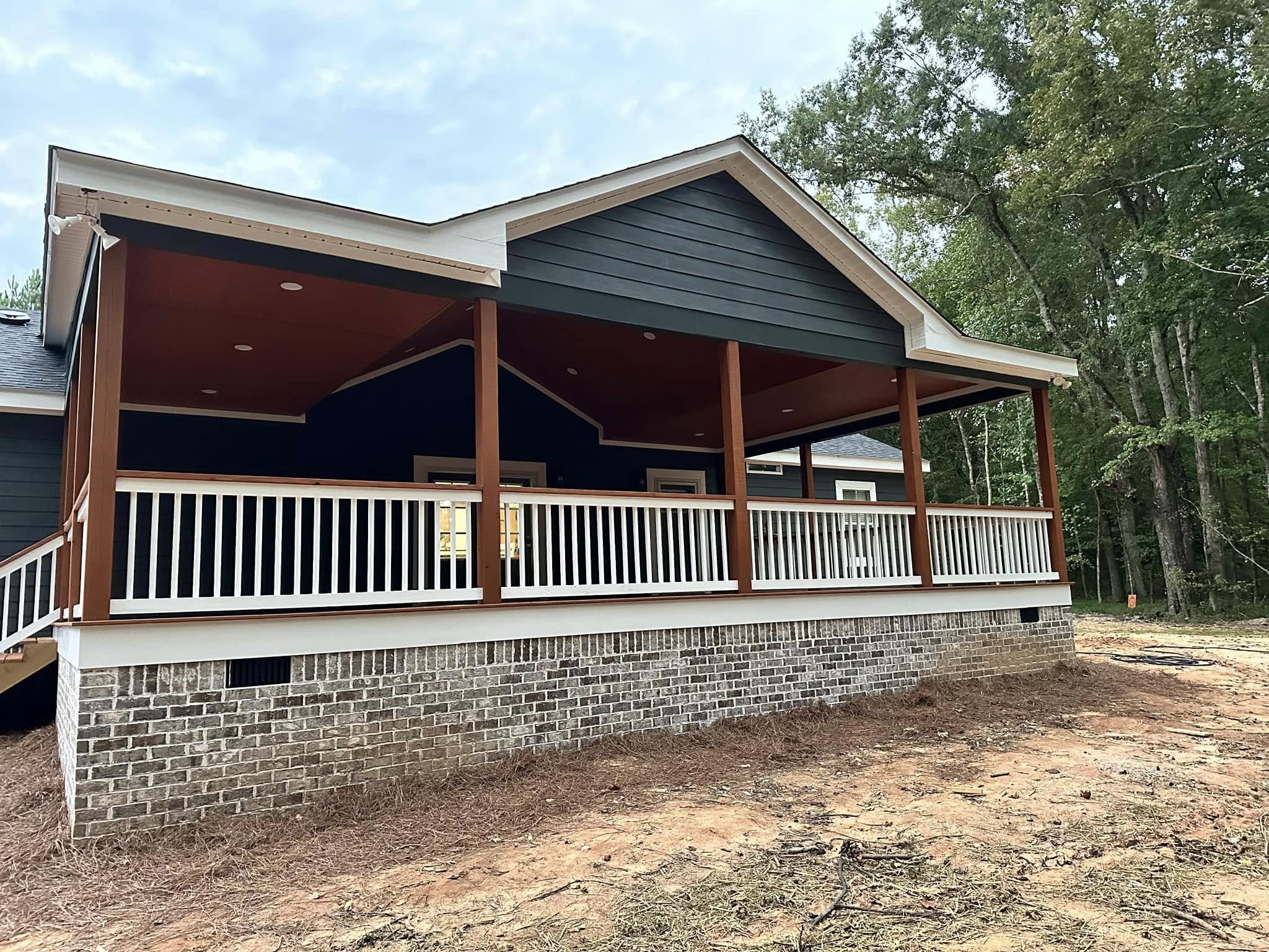A newly built house with a dark exterior, large covered porch with white railing, and brick foundation. Surrounded by trees, it conveys a serene, rustic vibe.
