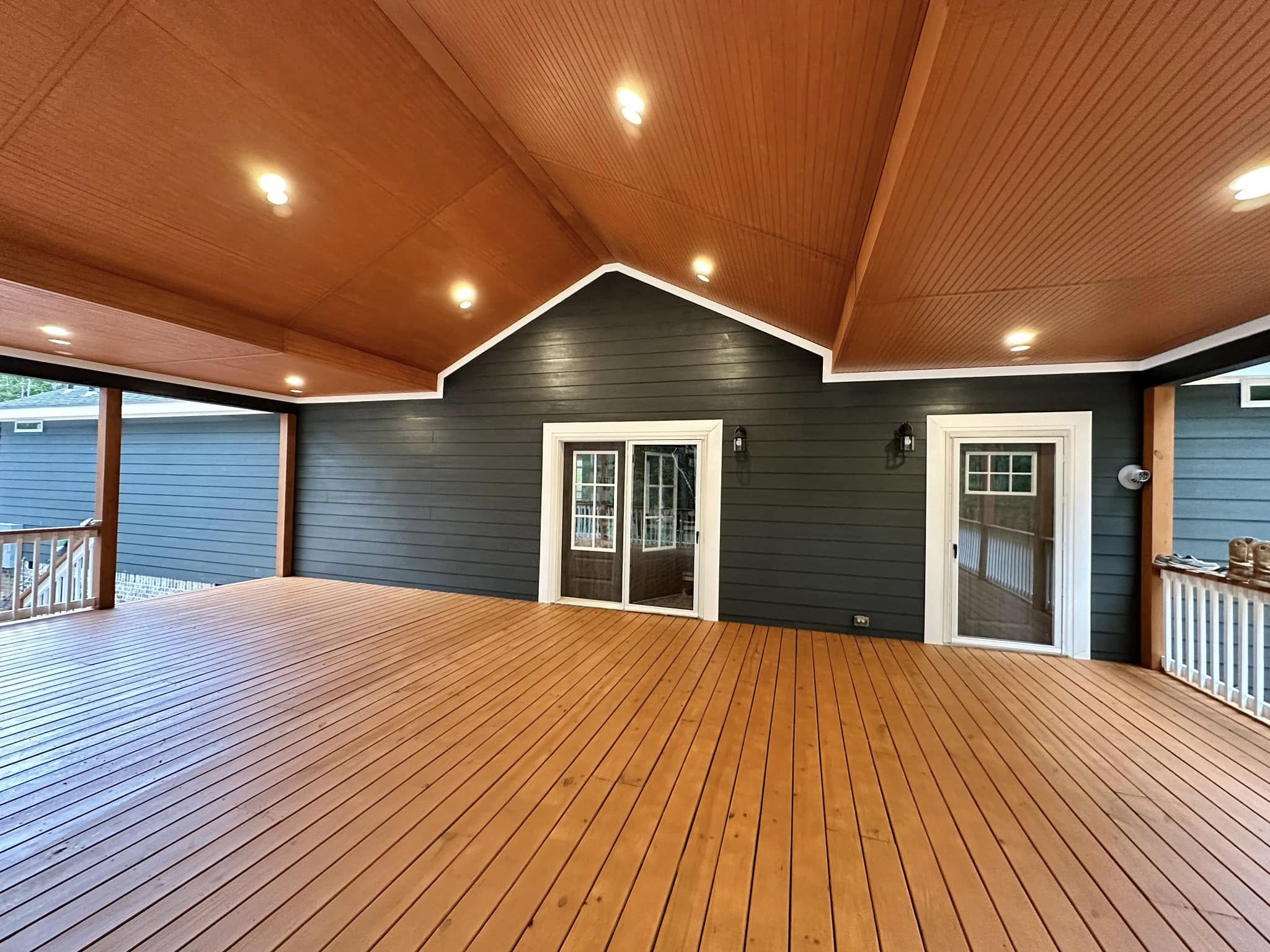 Covered wooden patio with a warm brown ceiling and floor. Dark blue siding wall features two white-framed doors. Recessed lighting creates a cozy ambiance.
