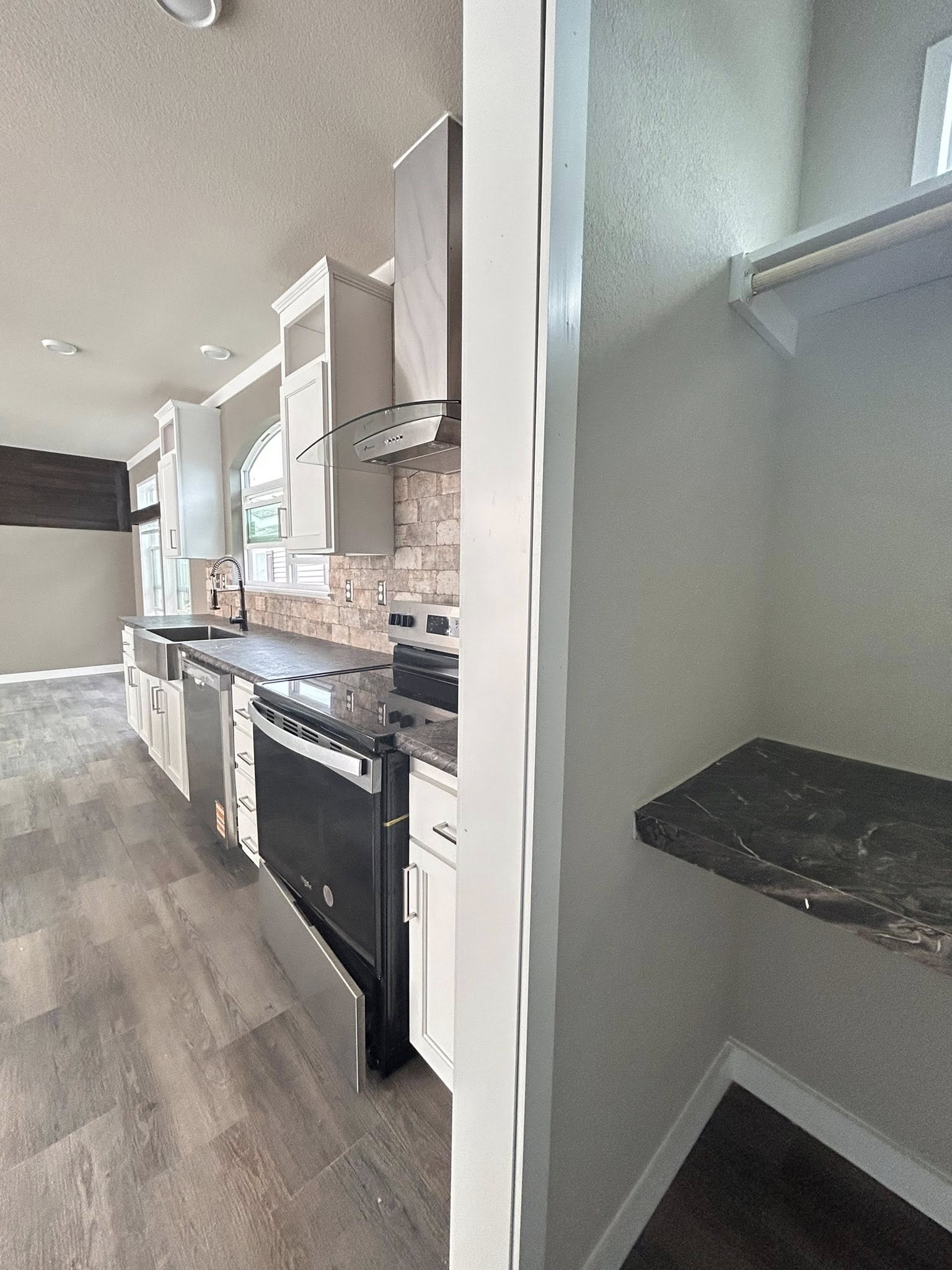 Modern kitchen with white cabinets, stainless steel appliances, and dark countertops. Adjacent room has a narrow shelf and light-colored walls.